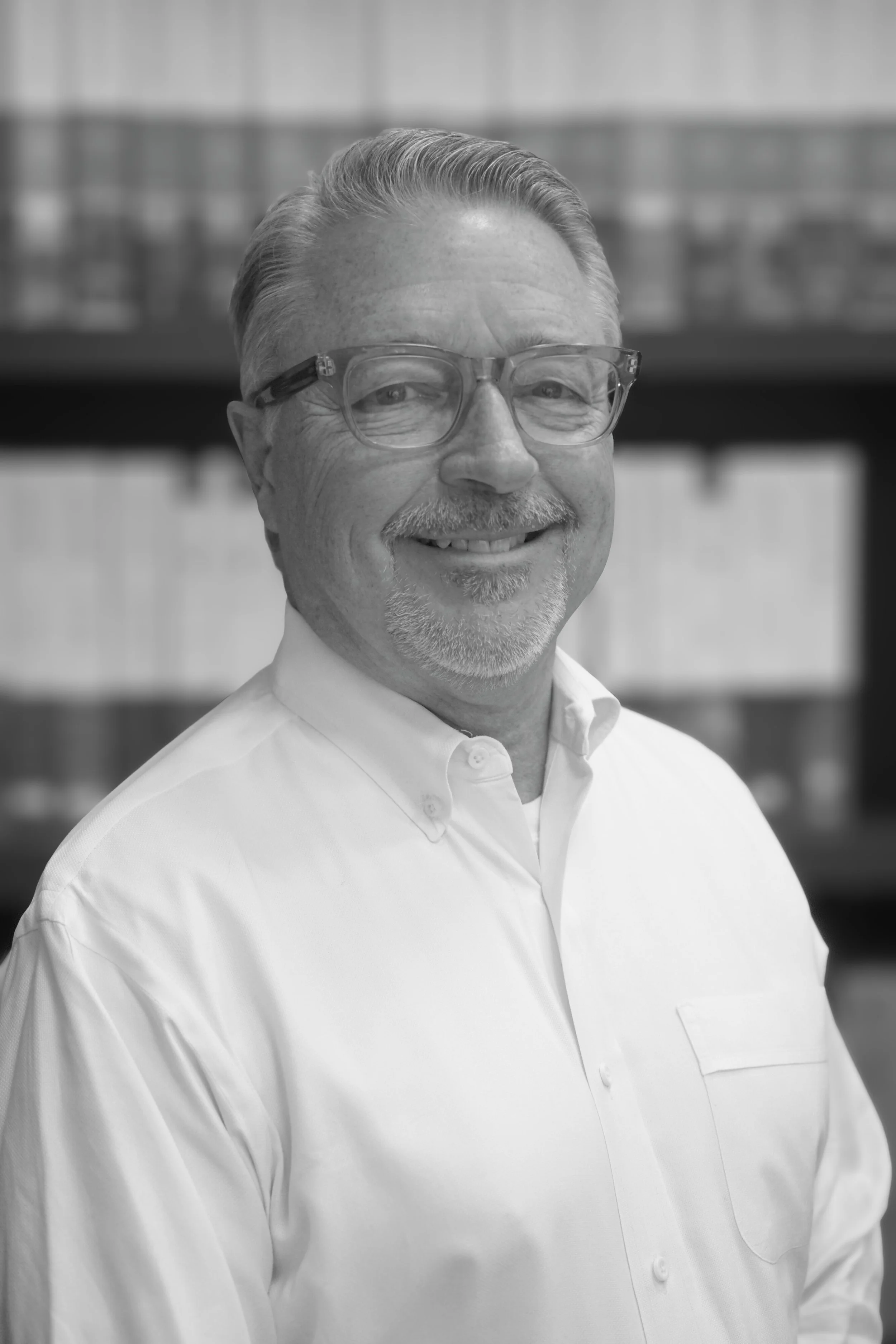 A smiling middle-aged man with glasses, short gray hair, and a mustache in a white shirt, standing against a blurred background.