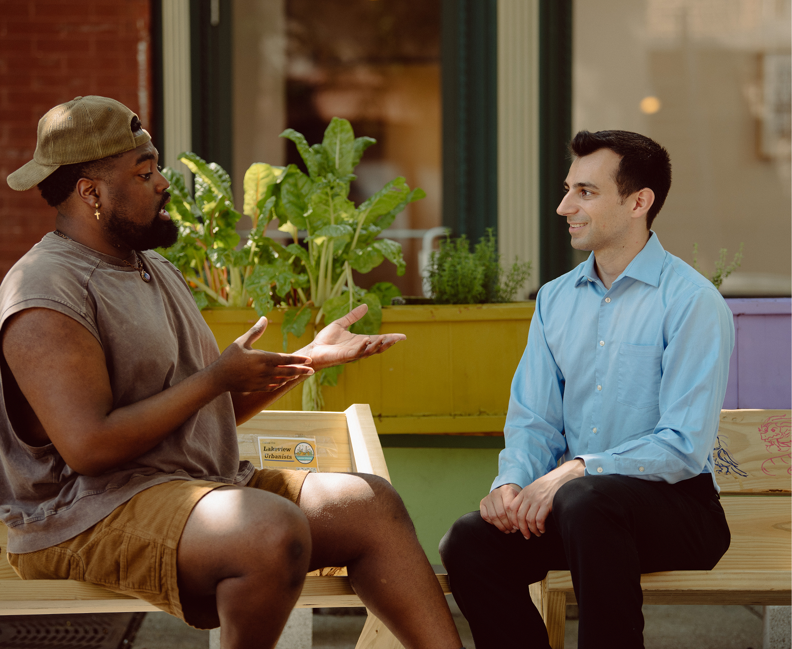 Two men sit across from each other having a conversation outdoors, with plants and a yellow planter behind them.