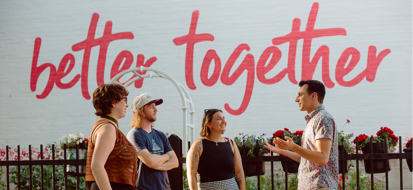 Four people standing outside near a fence with colorful flowers, engaged in conversation, with a large mural background that reads 'better together' in red letters.