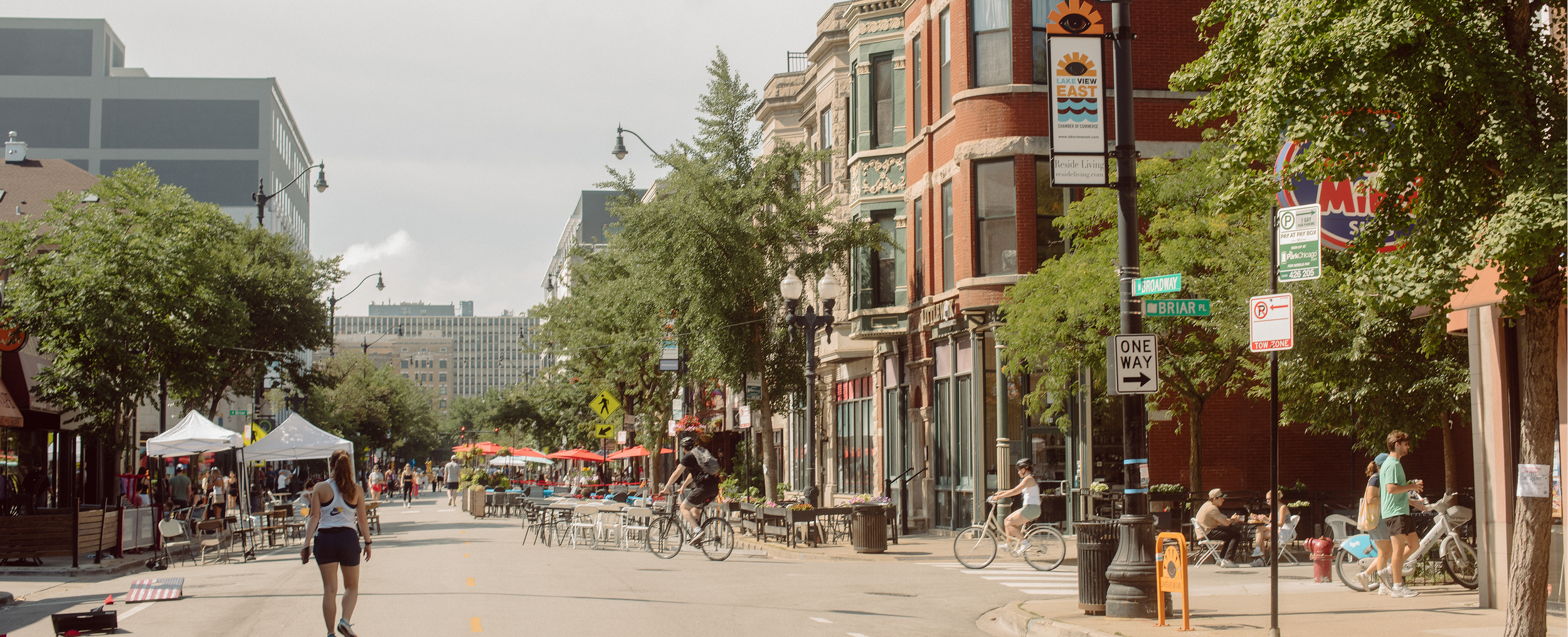 Busy city street with cyclists and pedestrians, outdoor seating, trees, historic buildings, and street signs indicating street names and traffic direction.