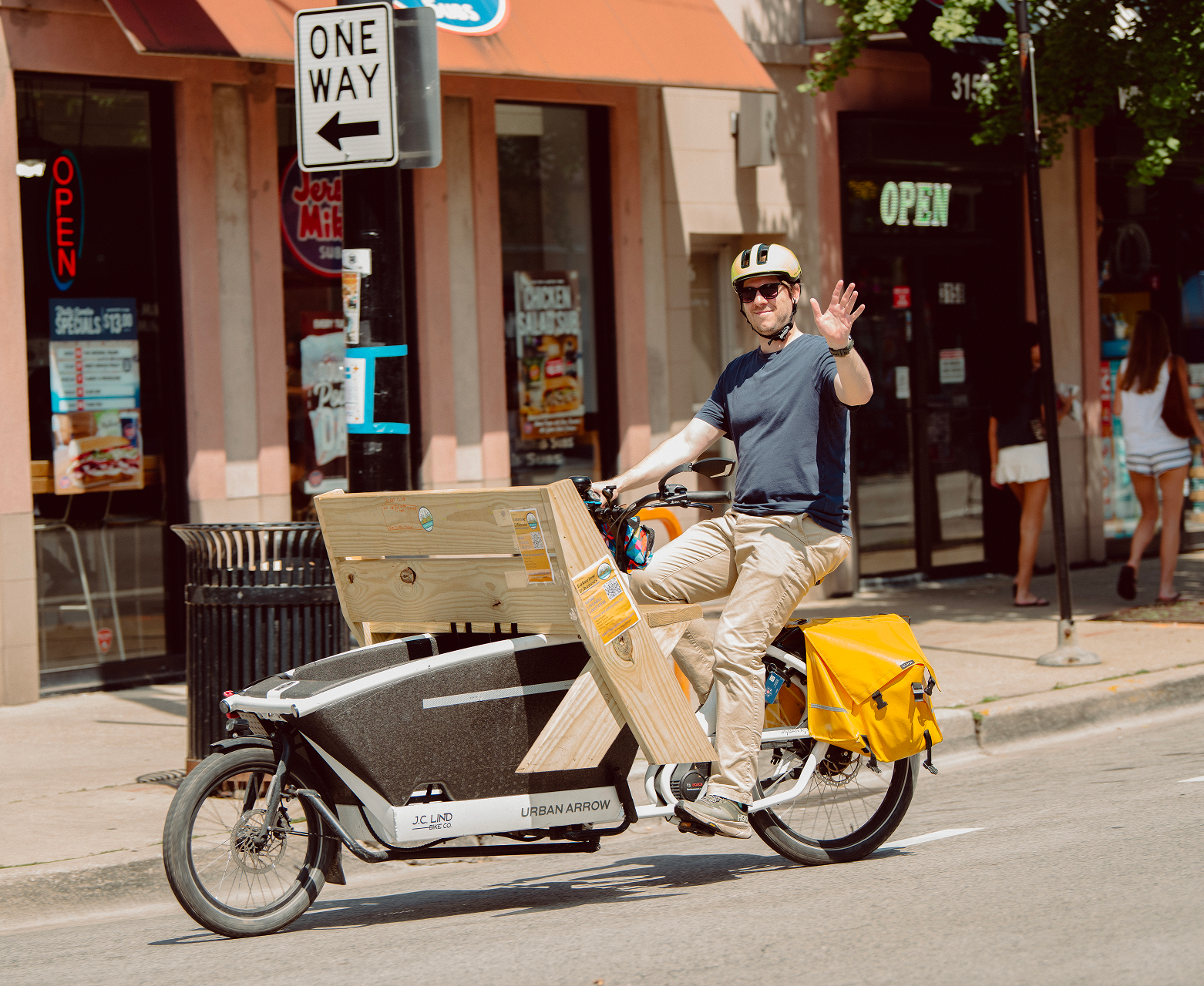 A man riding an electric bike with a cargo box on a city street, waving and smiling, with stores and pedestrians in the background.