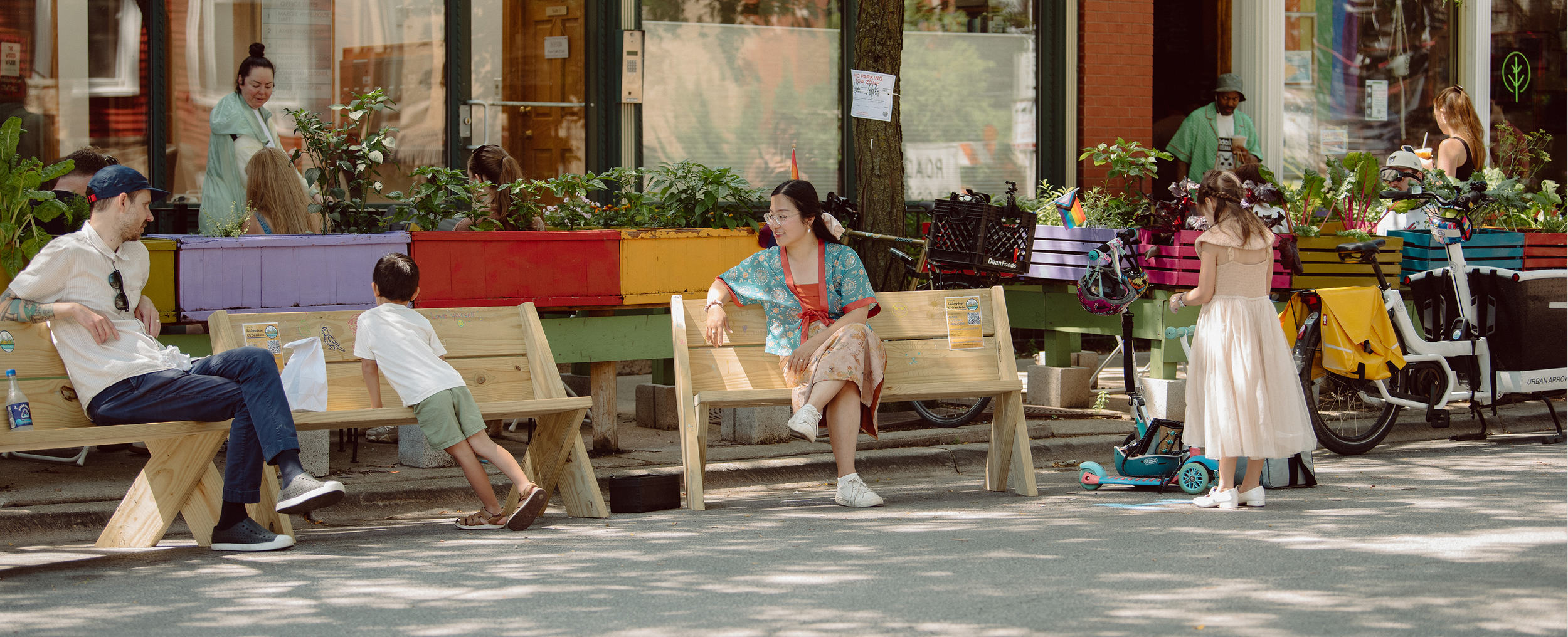 People sitting and standing on a sidewalk in front of a storefront with colorful planter boxes, bicycles, and green trees, enjoying a sunny day.