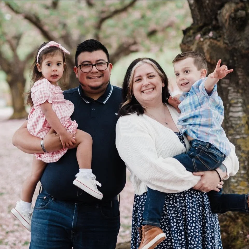 A happy family of four standing outdoors under a large tree, smiling at the camera, with the parents holding their young children in their arms.
