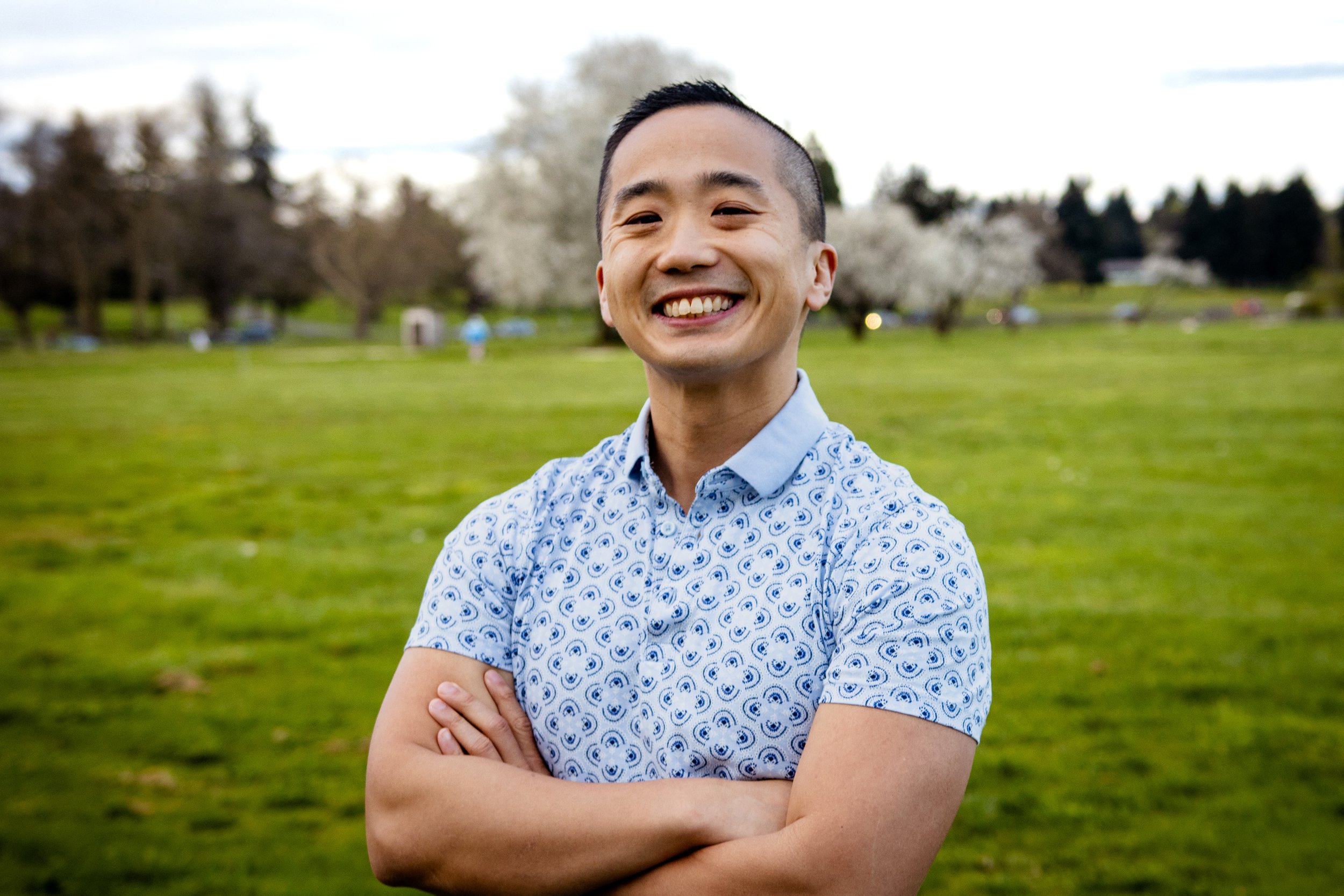 A young man smiling outdoors in a park with trees and grass in the background, arms crossed, wearing a patterned short-sleeve shirt.
