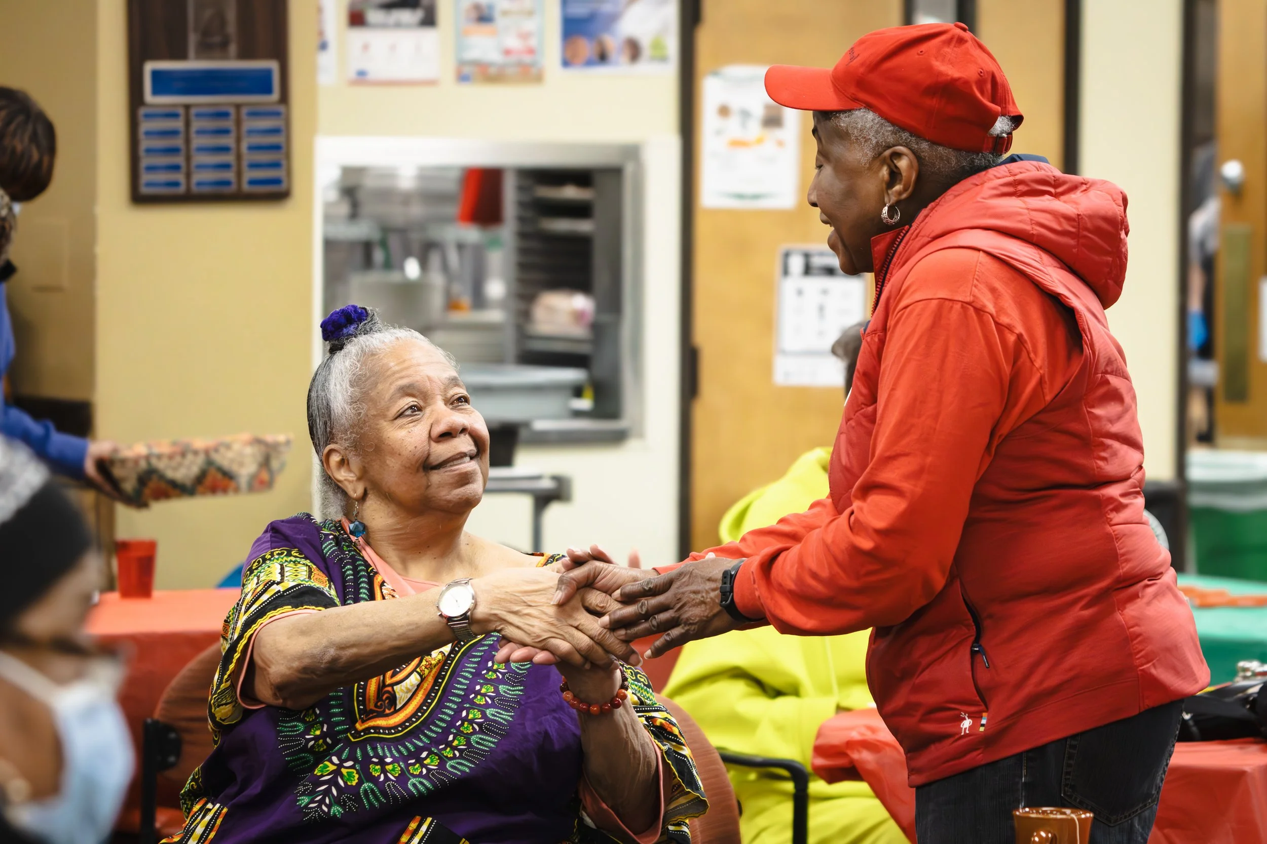 An elderly woman with grey hair and colorful traditional African clothing holding hands and smiling at a woman in a red jacket and cap, who is leaning over and smiling at her during a social gathering.