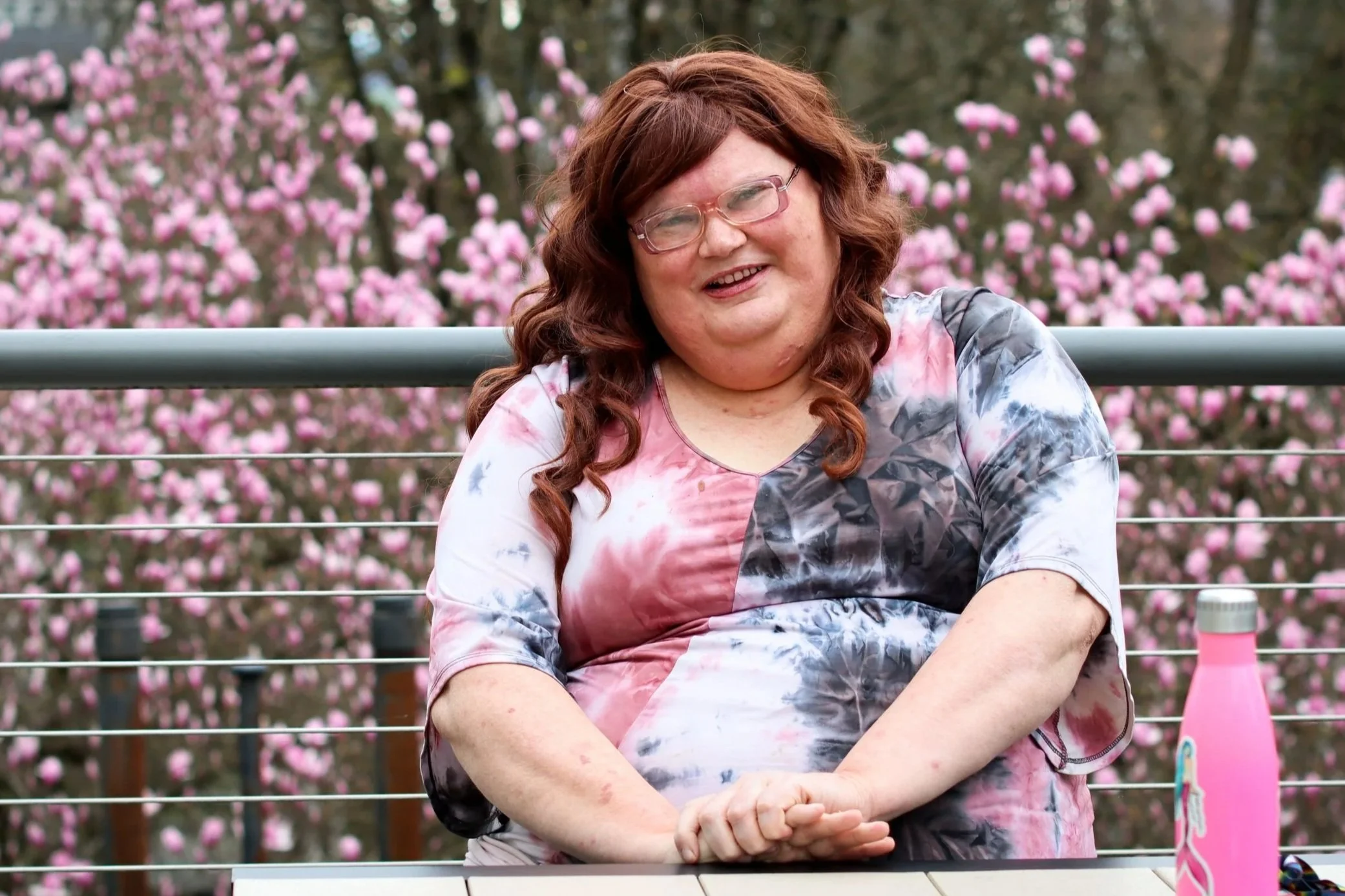 A woman with curly red hair and glasses smiling, sitting outdoors in front of pink flowering trees, wearing a tie-dye shirt.