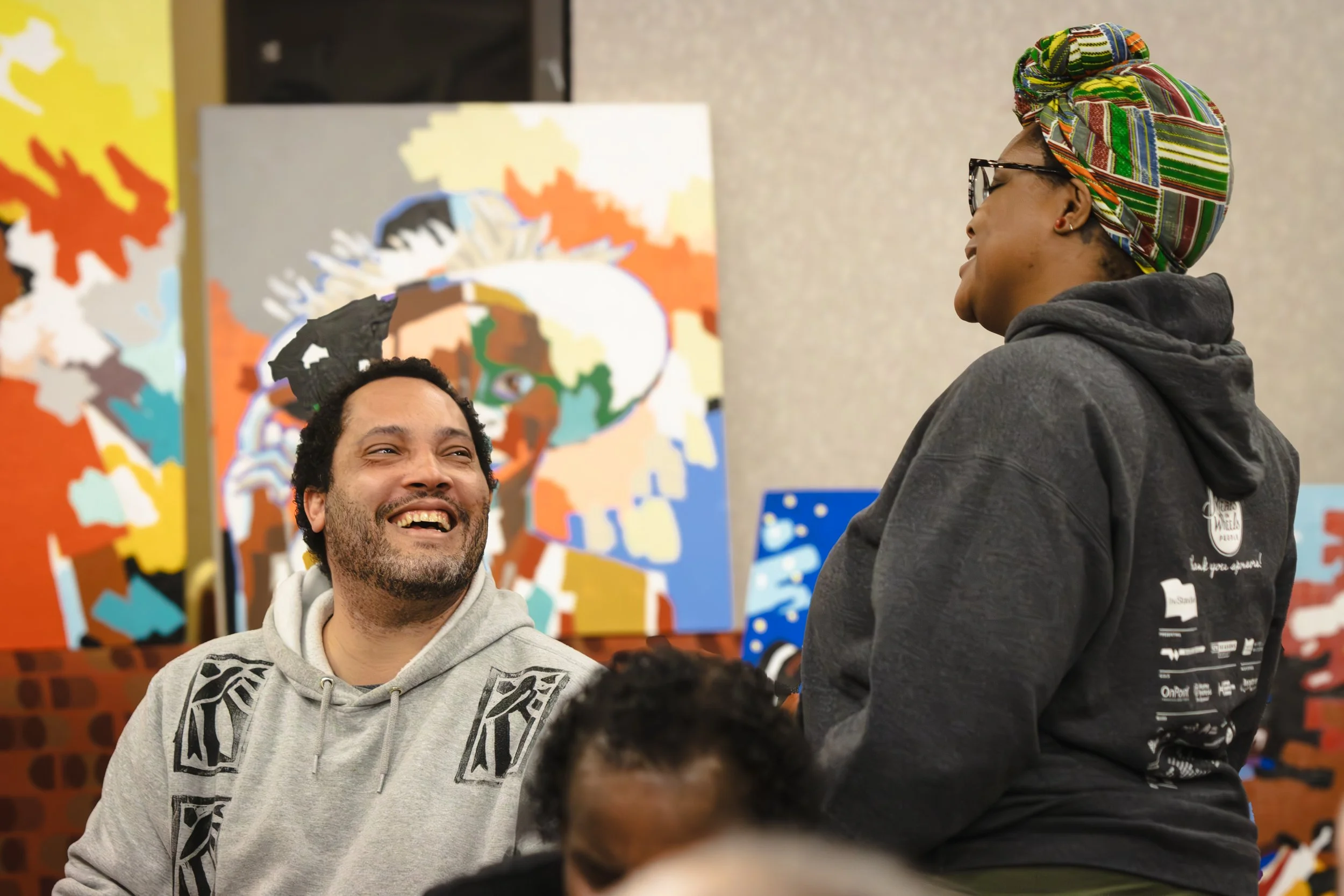 Two people smiling and conversing in front of colorful artwork, one wearing a gray hoodie and the other wearing a multicolored headwrap and black hoodie.