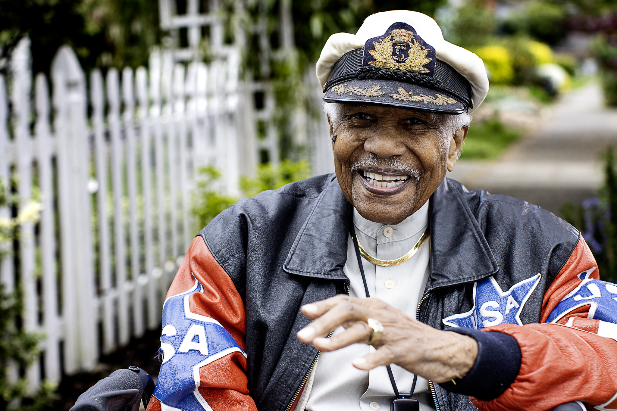An elderly man wearing a black and white captains hat with gold embroidery, a leather jacket decorated with a large colored patch, and gold jewelry, smiling outdoors.