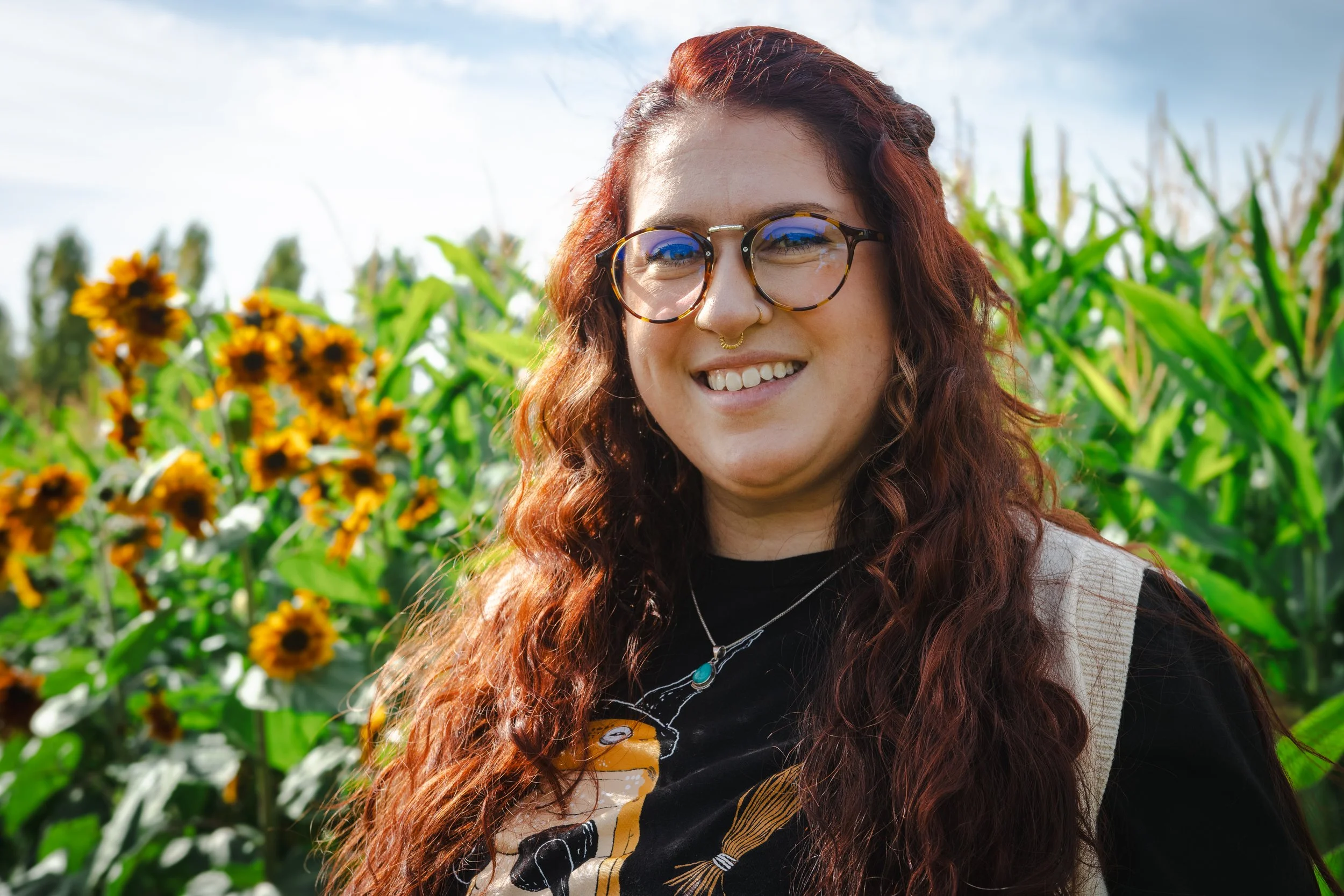 A smiling woman with long curly red hair and glasses, standing outdoors among tall green plants and sunflowers, with a blue sky in the background.