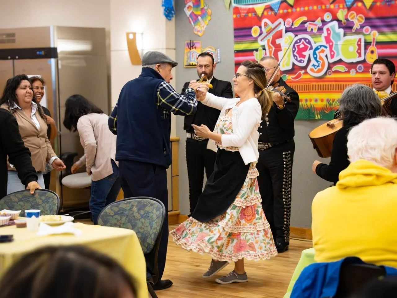 People dancing and playing music at a festive celebration, with a colorful banner that says "Fiesta" in the background.