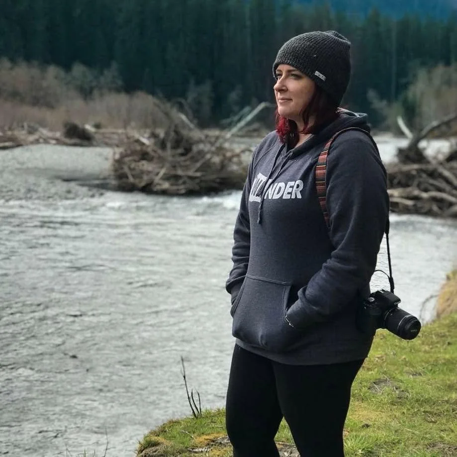 Woman standing near a river with trees and driftwood in the background, wearing a gray hoodie, black beanie, and carrying a camera.
