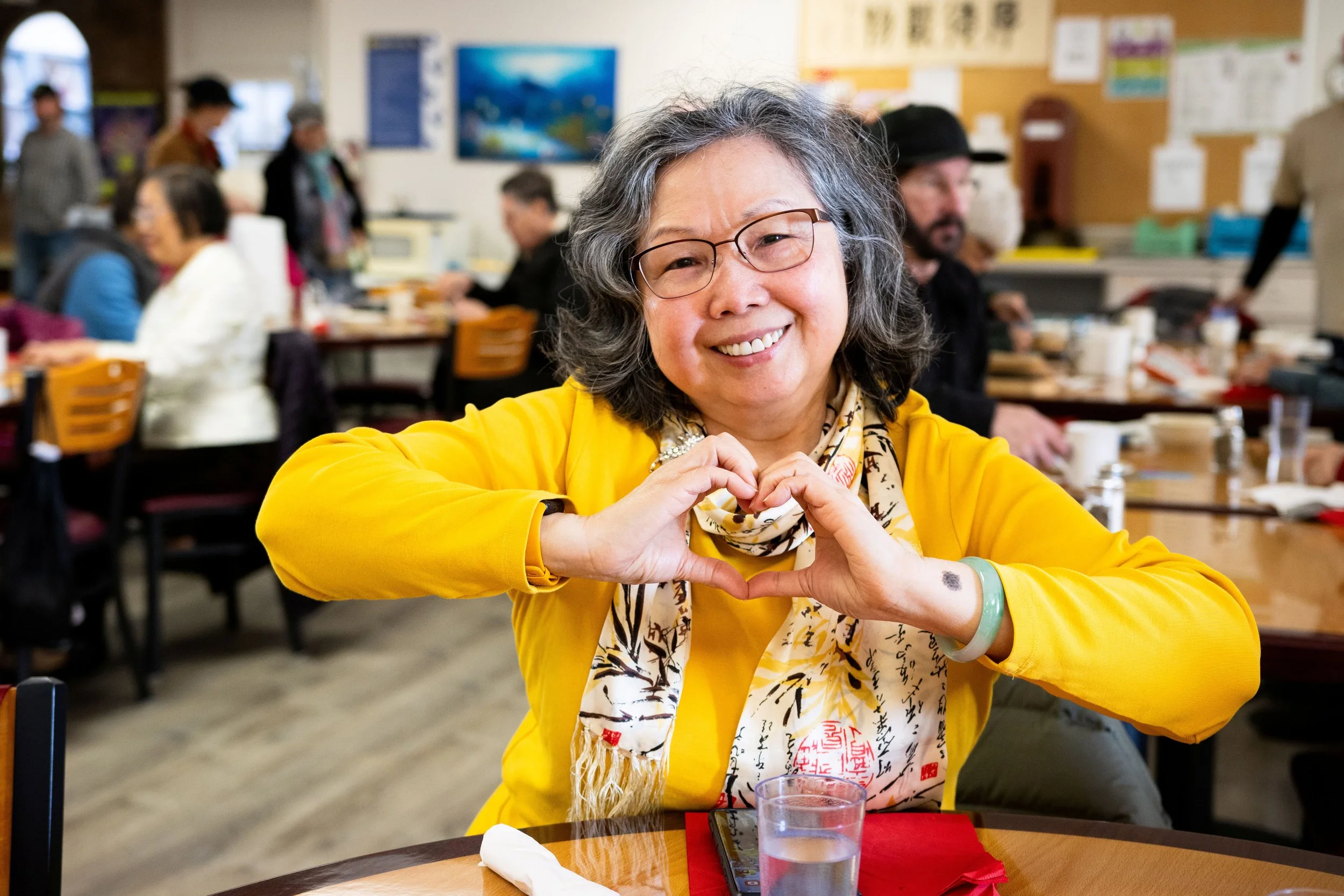 A woman in a yellow shirt and glasses makes a heart shape with her hands while sitting at a table. She is smiling and appears happy. There are other people in the background dining and socializing.