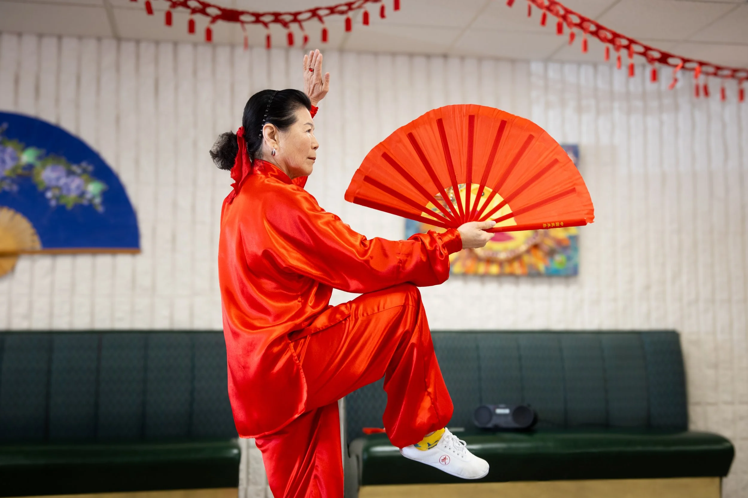 A woman dressed in red traditional attire practicing fan dance indoors, holding a red fan, with decorations and paintings in the background.