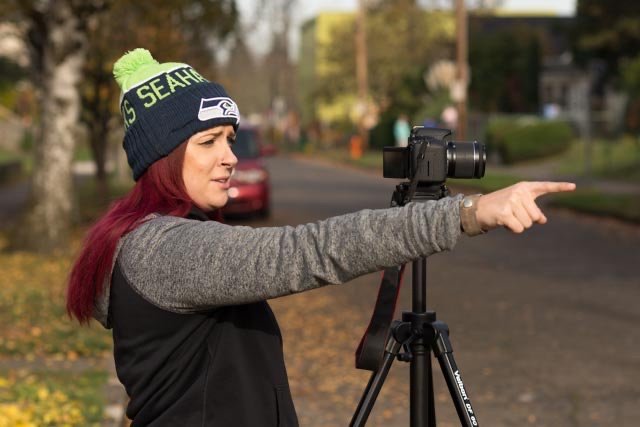 A woman with red hair wearing a Seahawks beanie and a gray and black jacket, pointing at a camera on a tripod outdoors in a neighborhood with trees and houses.