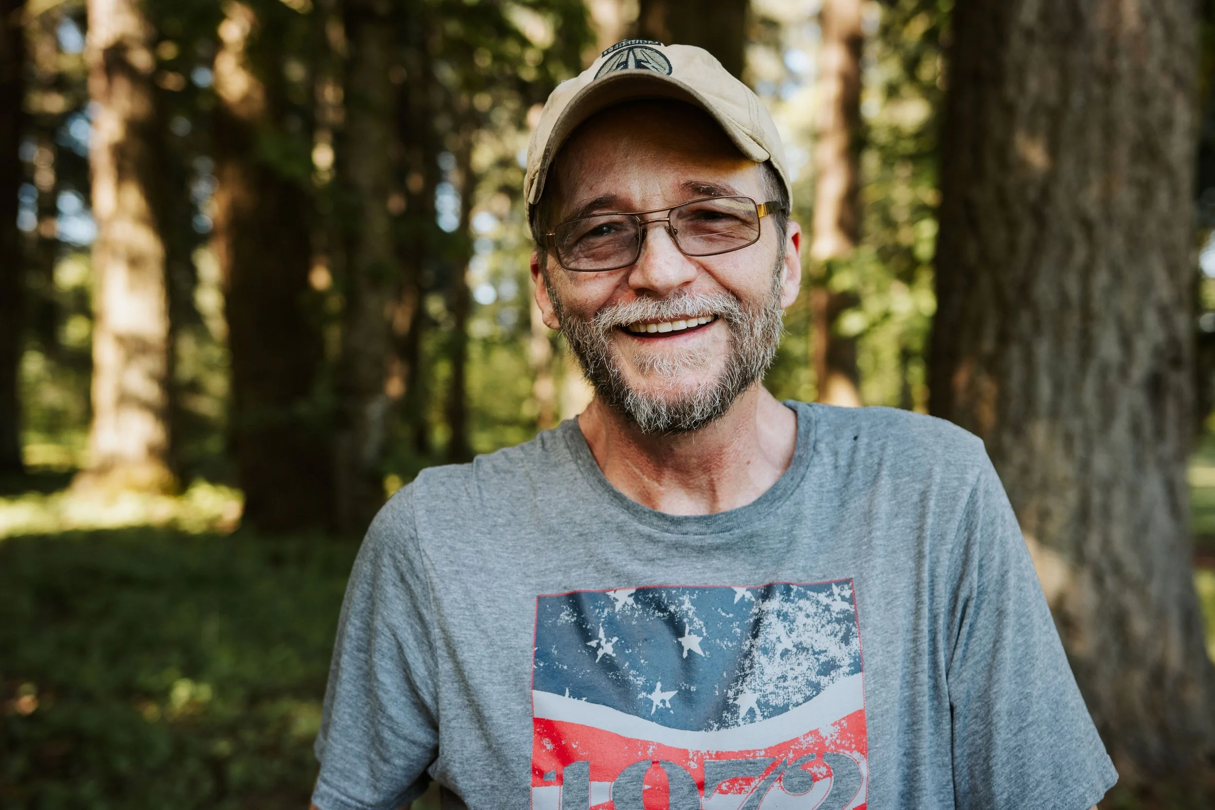 A smiling middle-aged man with a beard and glasses, wearing a beige cap and a gray T-shirt with a red, white, and blue graphic, standing outdoors in a forest with tall trees and green foliage in the background.