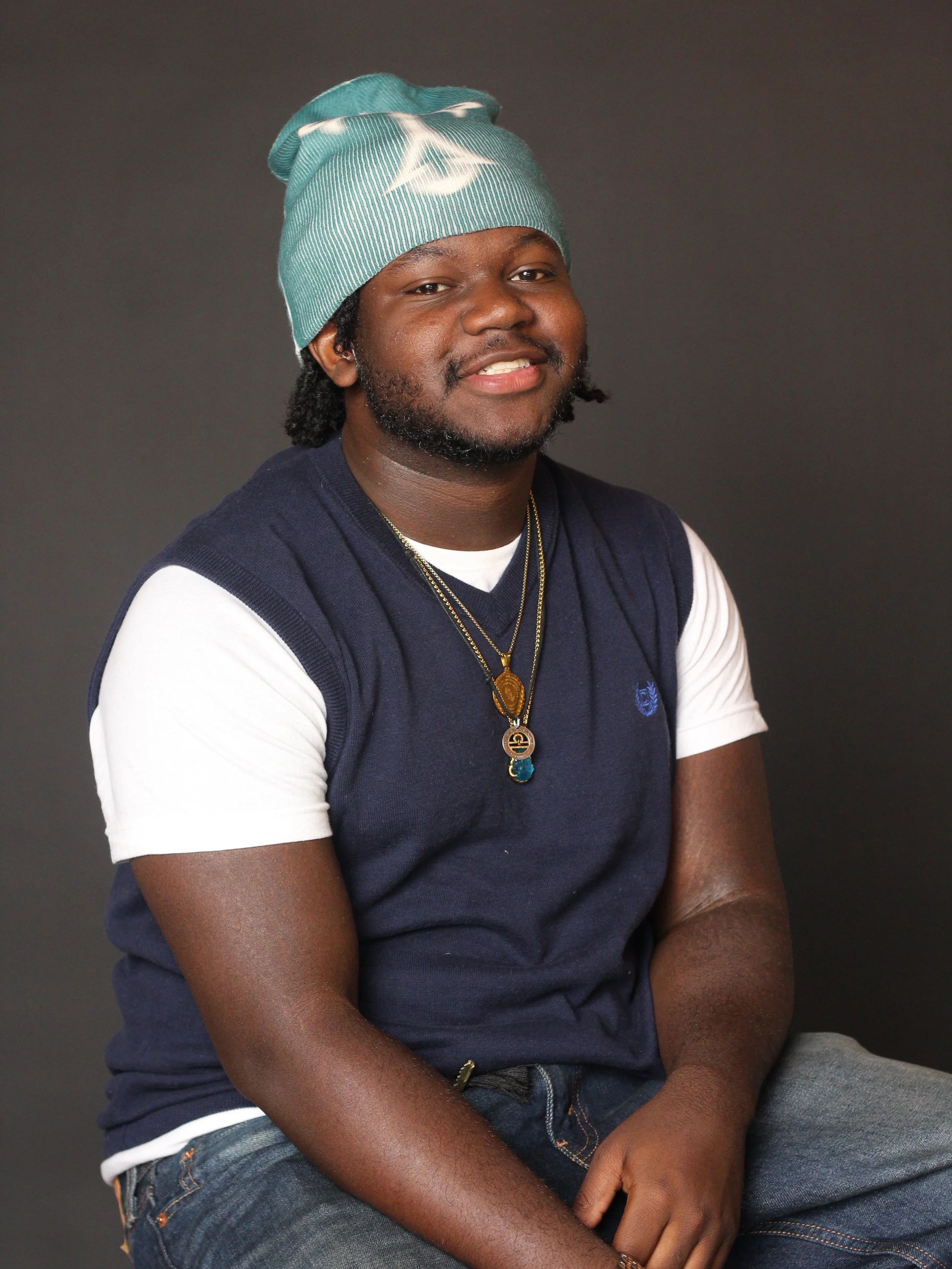 Portrait of a smiling man with a beard, wearing a teal beanie, layered with a navy vest over a white t-shirt, and layered necklaces, sitting against a dark background.
