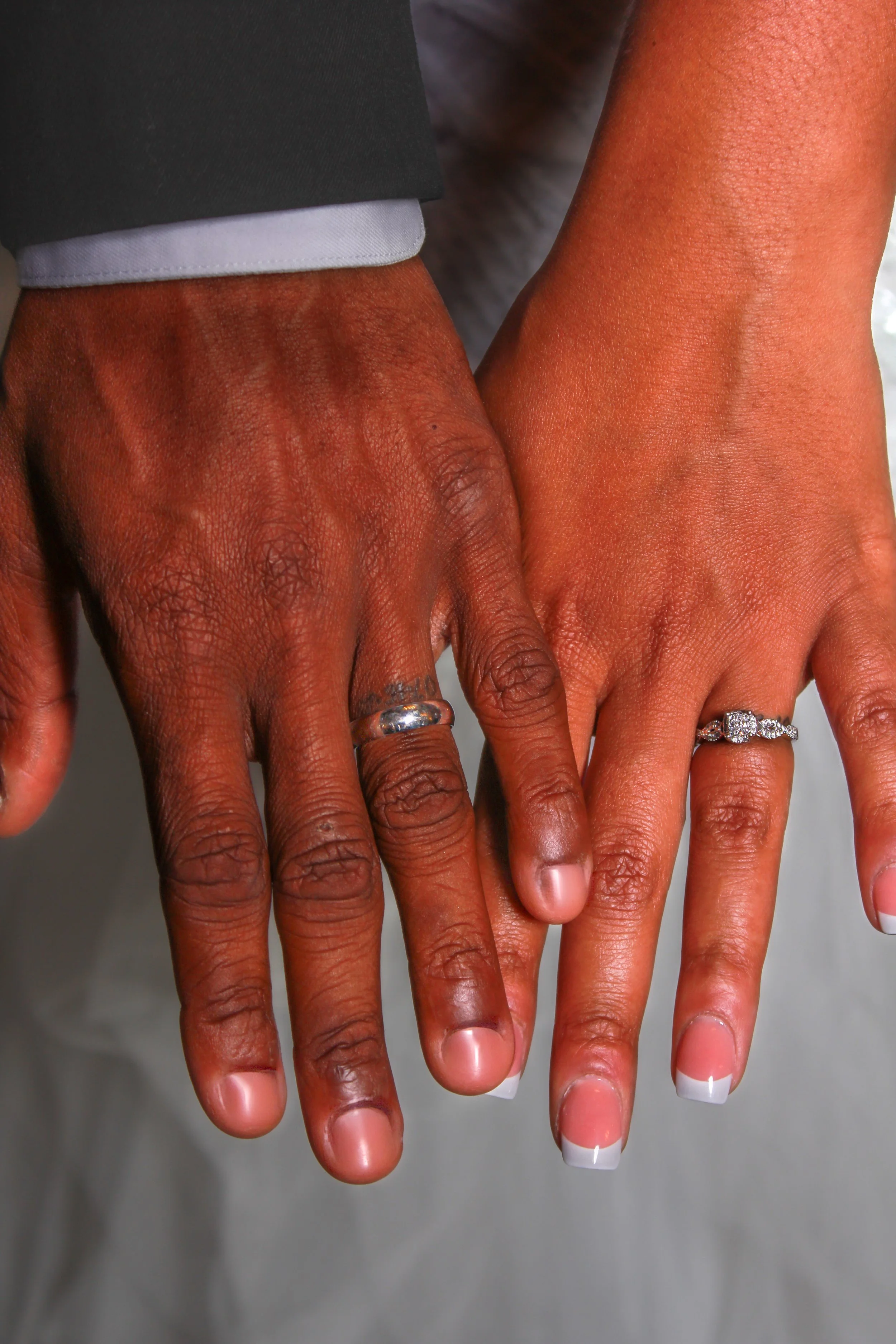 Two hands with different skin tones, one with darker skin and one with lighter skin, both wearing rings, touching gently with fingers extended, against a gray background.
