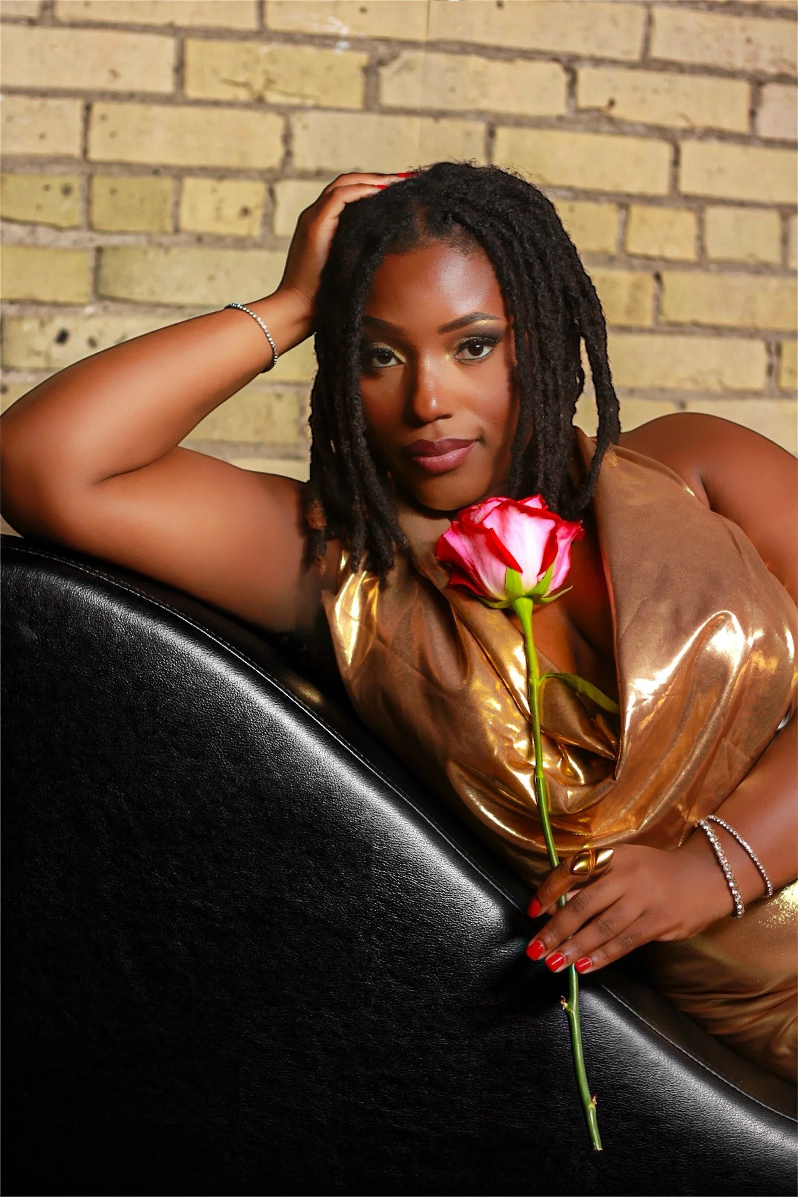 A woman with dreadlocks reclining against a black surface, holding a pink and white rose, wearing a shiny brown top, and looking into the camera with a confident expression in front of a brick wall.
