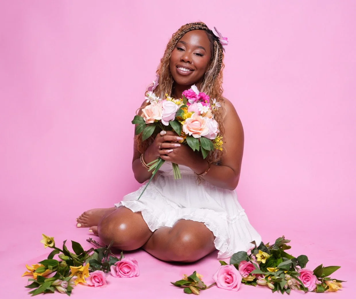 A woman with long, blonde, braided hair, sitting on her knees on a pink background, holding a bouquet of pink and white flowers. She wears a white dress and has a pink flower in her hair. Pink roses and yellow flowers are arranged on the floor around