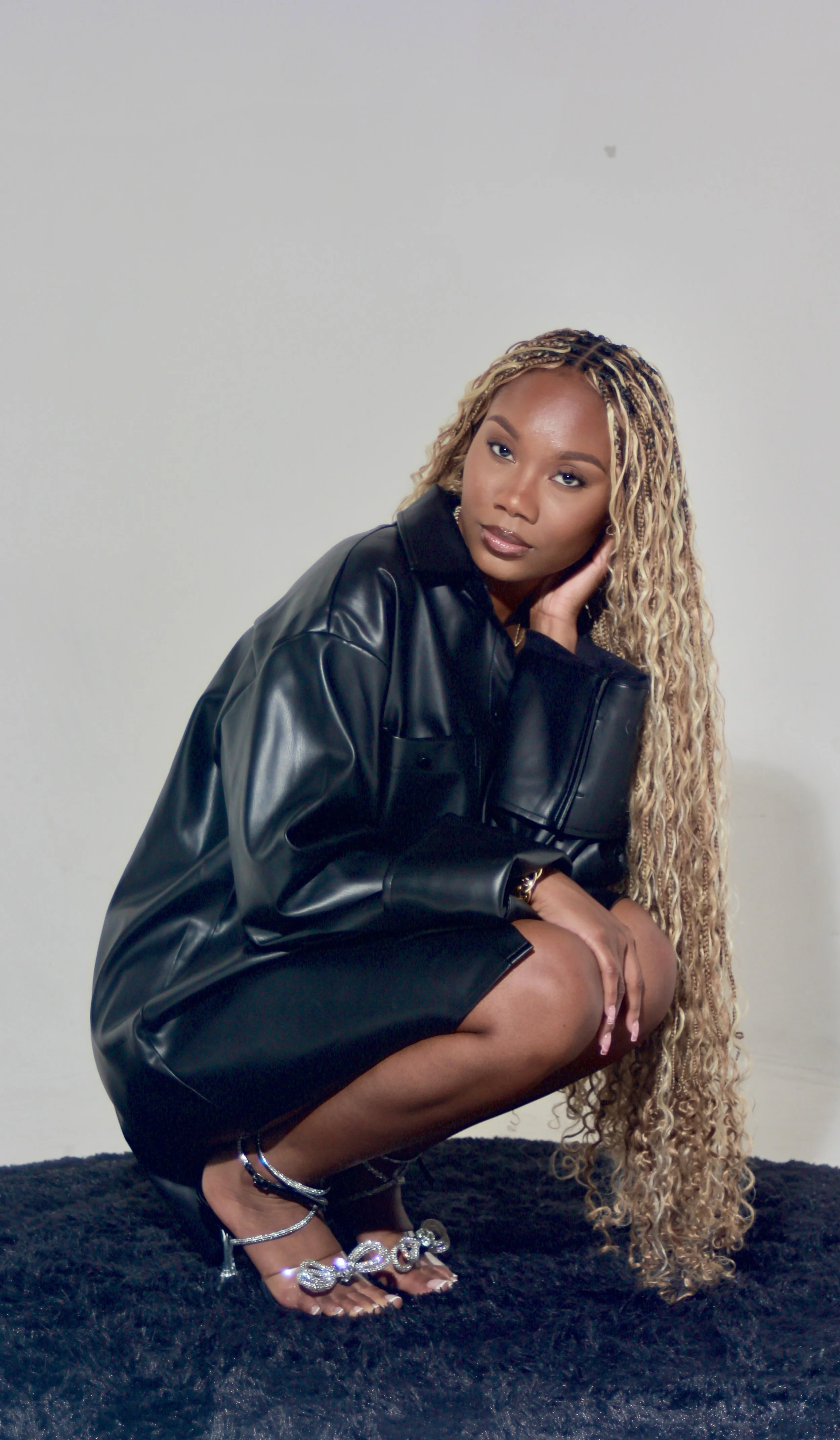 A woman with long, curly blonde hair squatting on a soft black rug. She is wearing a black leather jacket and black shorts, with strappy high-heeled sandals decorated with large rhinestones. The woman has a calm expression, gazing directly at the cam
