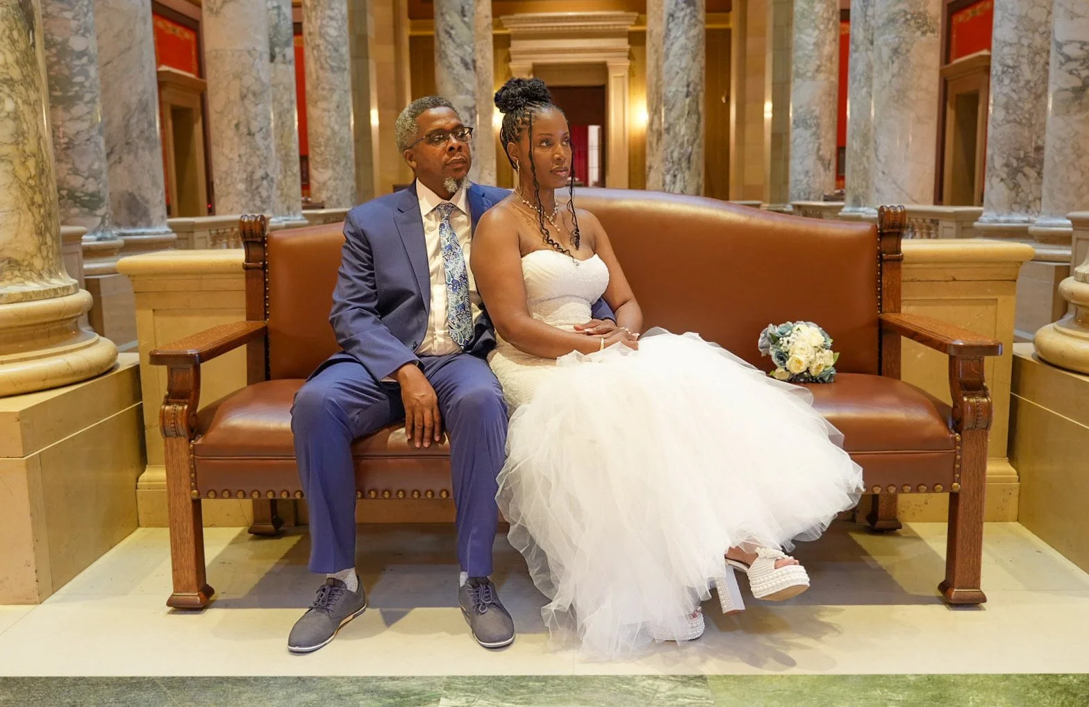 A man and a woman sitting on a brown bench in an elegant hall with marble columns. The woman wears a white wedding dress with a floral bouquet beside her, and the man wears a blue suit with a white shirt and patterned tie.