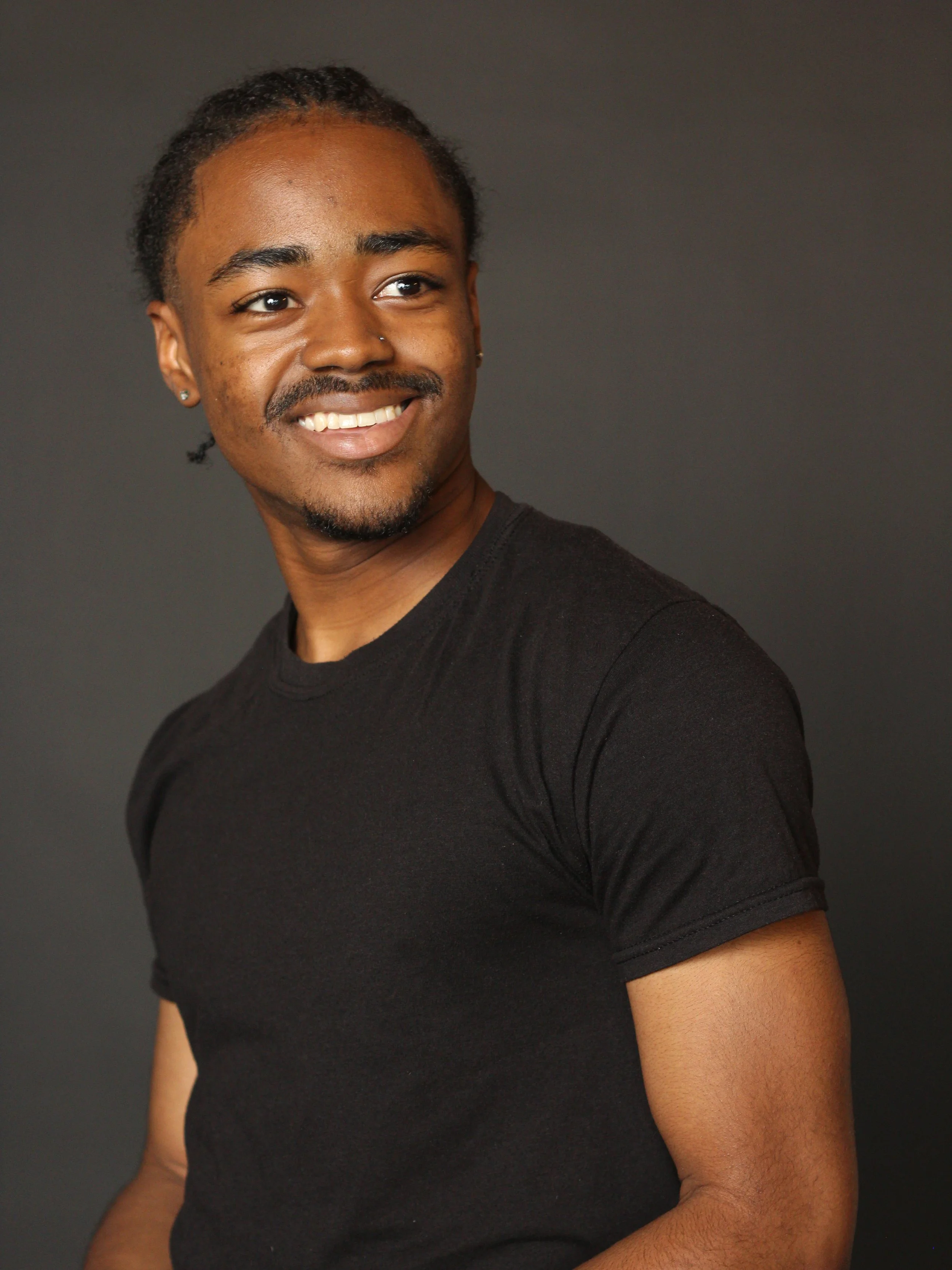 A young man with a mustache and goatee, smiling, wearing a black t-shirt, against a gray background.