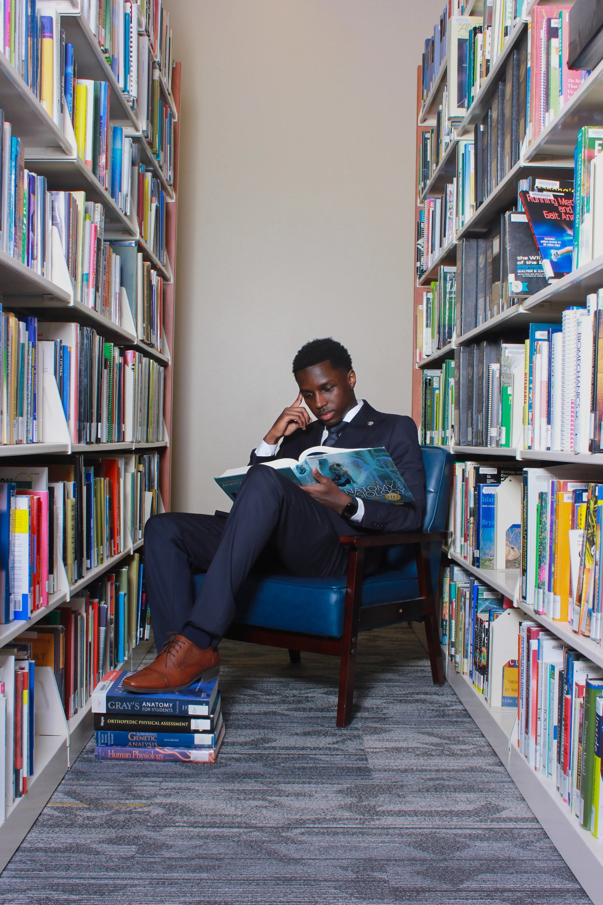 A young man in a suit sitting in a library between two shelves of books, reading a book, with more books stacked on the floor and a colorful carpet below him.