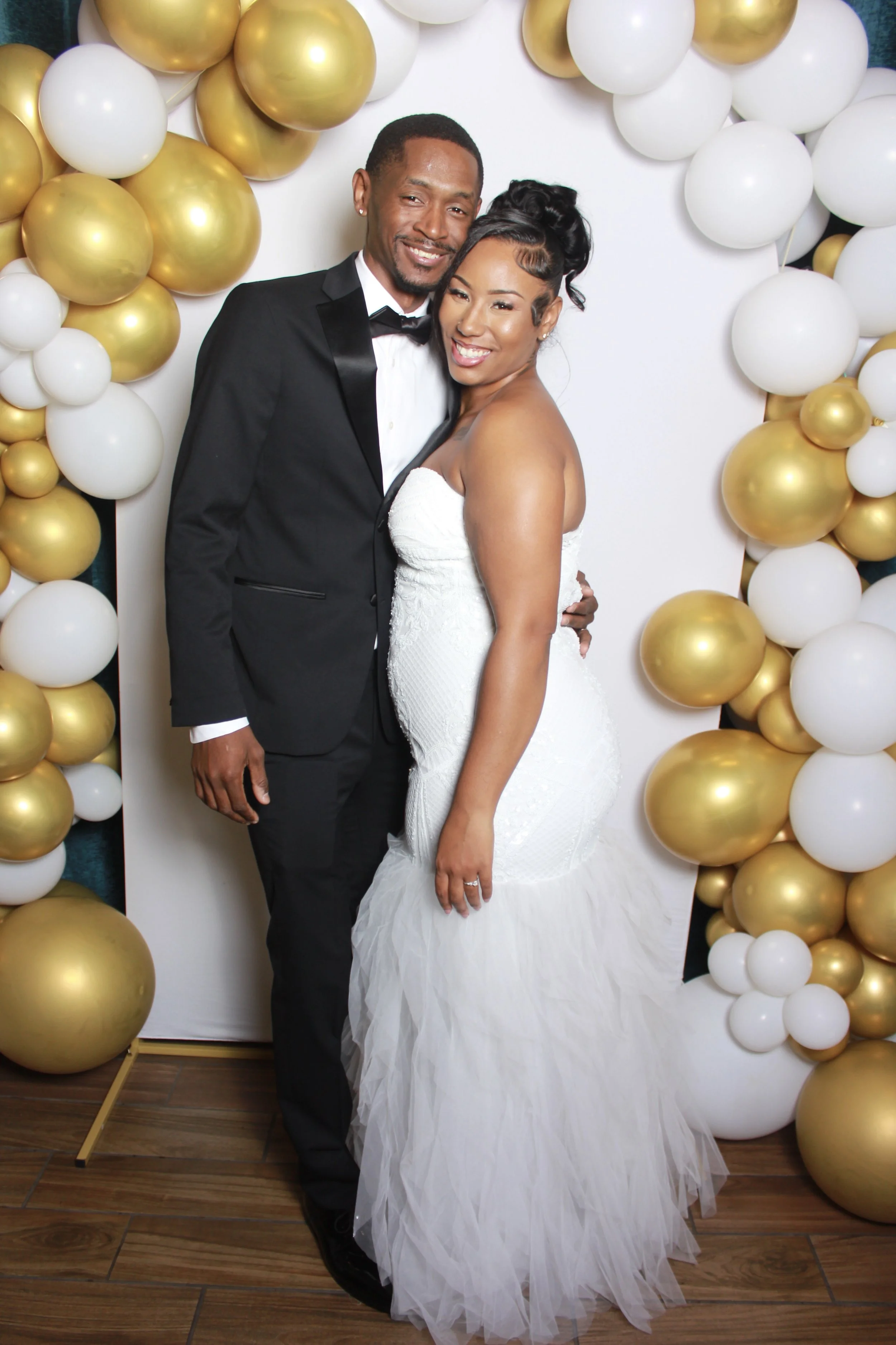 A newlywed couple smiling and posing together at a wedding reception, surrounded by a decorative arch of white and gold balloons.