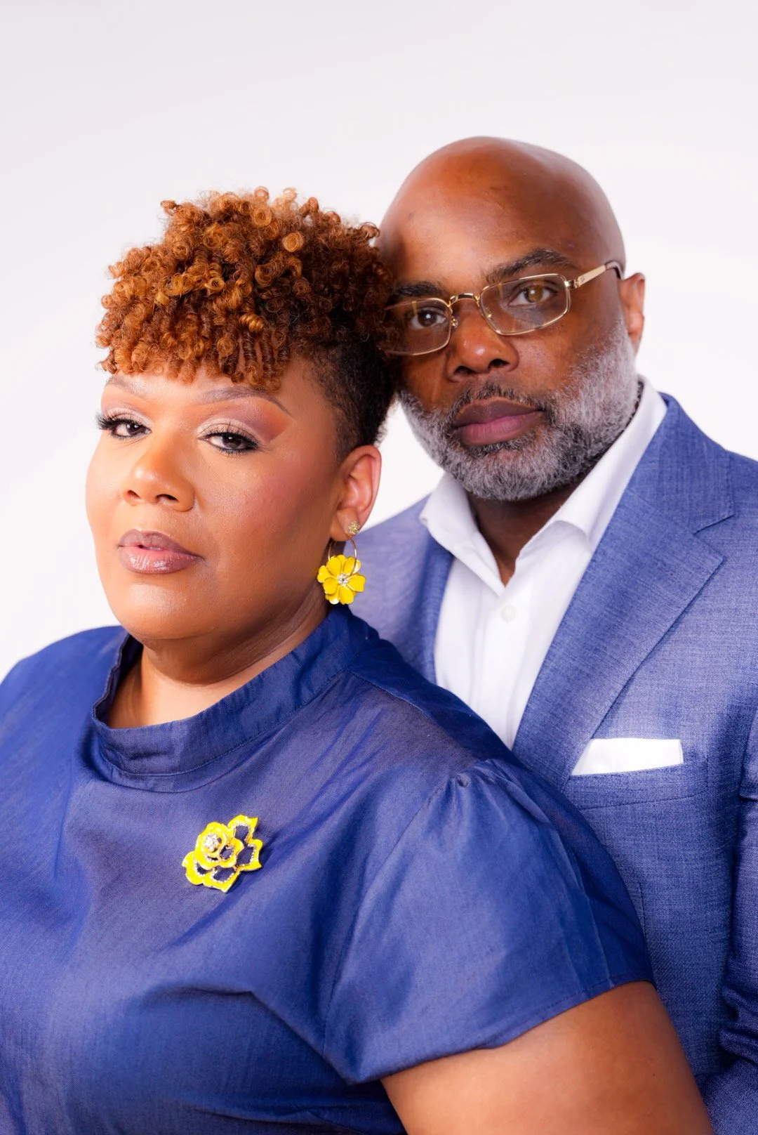 A serious-looking African American woman with short curly hair wearing a blue dress and yellow floral earrings, and an African American man with glasses, a beard, and bald head wearing a blue suit and white shirt, posing together against a white back
