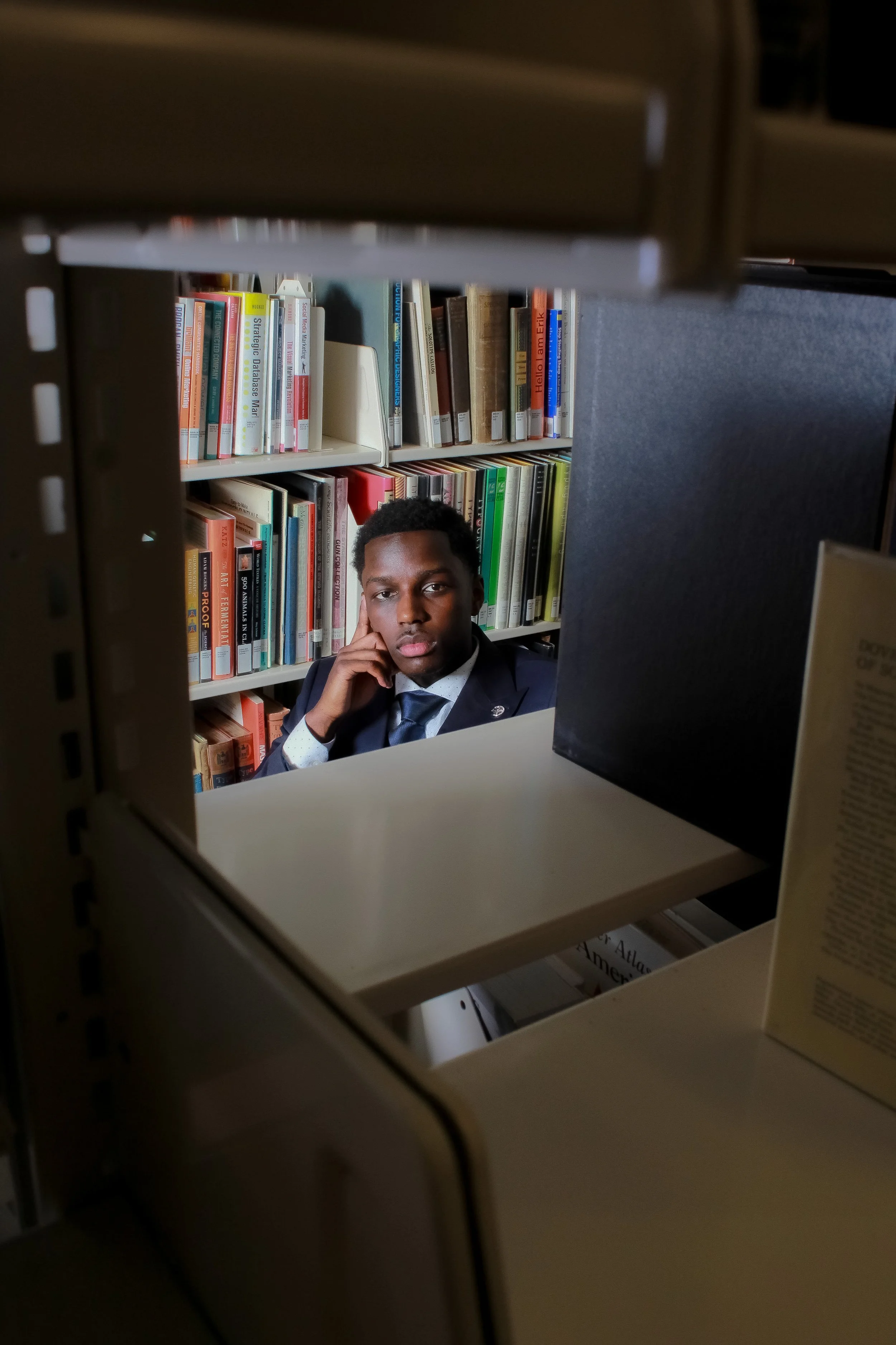 A young man in a suit and tie sitting in a library, looking at the camera, with bookshelves filled with colorful books behind him, seen through a small gap in a bookshelf.