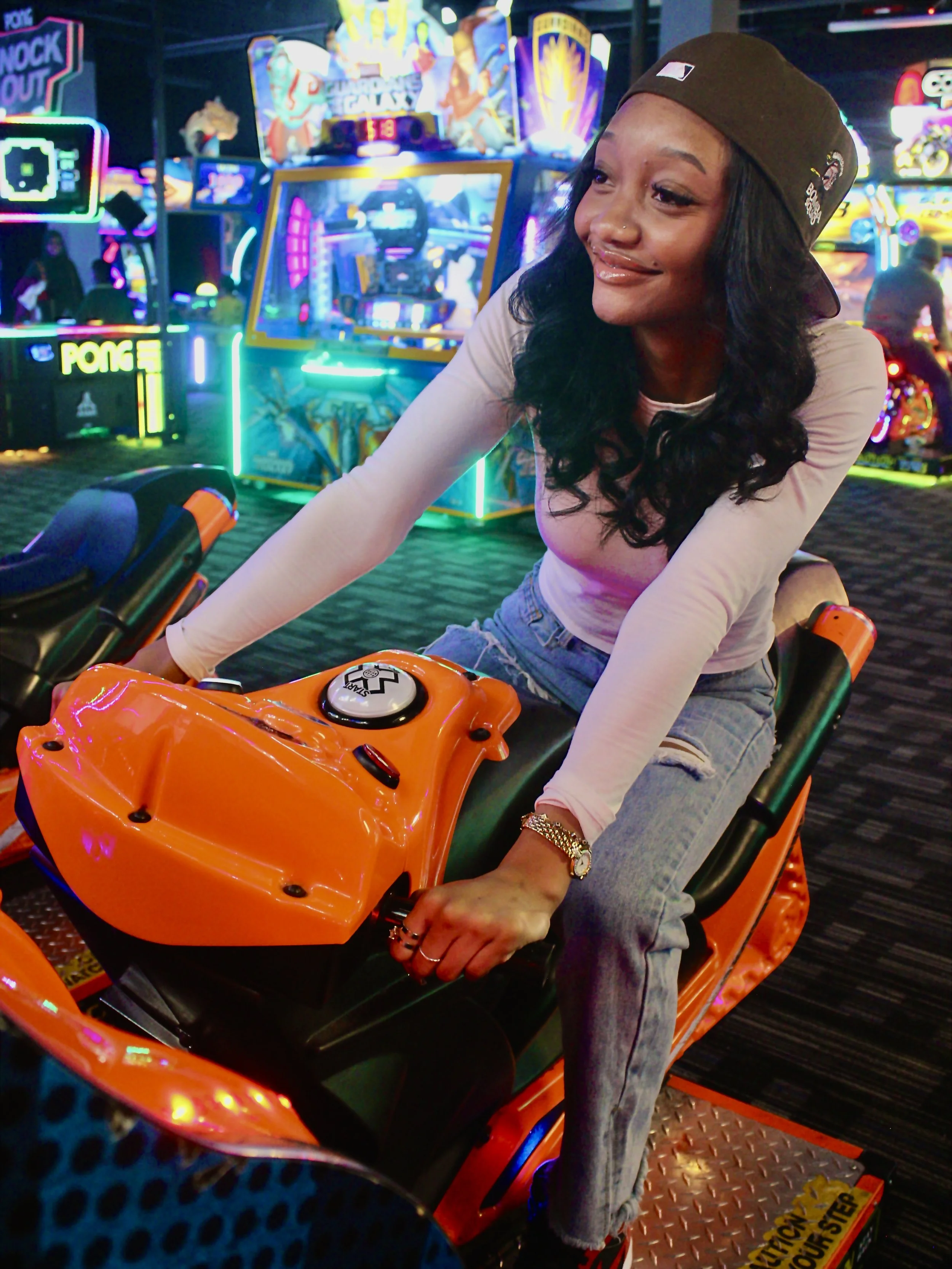 A woman with long black wavy hair and a nose piercing, wearing a brown baseball cap and a beige long-sleeve shirt, is riding an orange amusement park vehicle in a neon-lit arcade or amusement area.