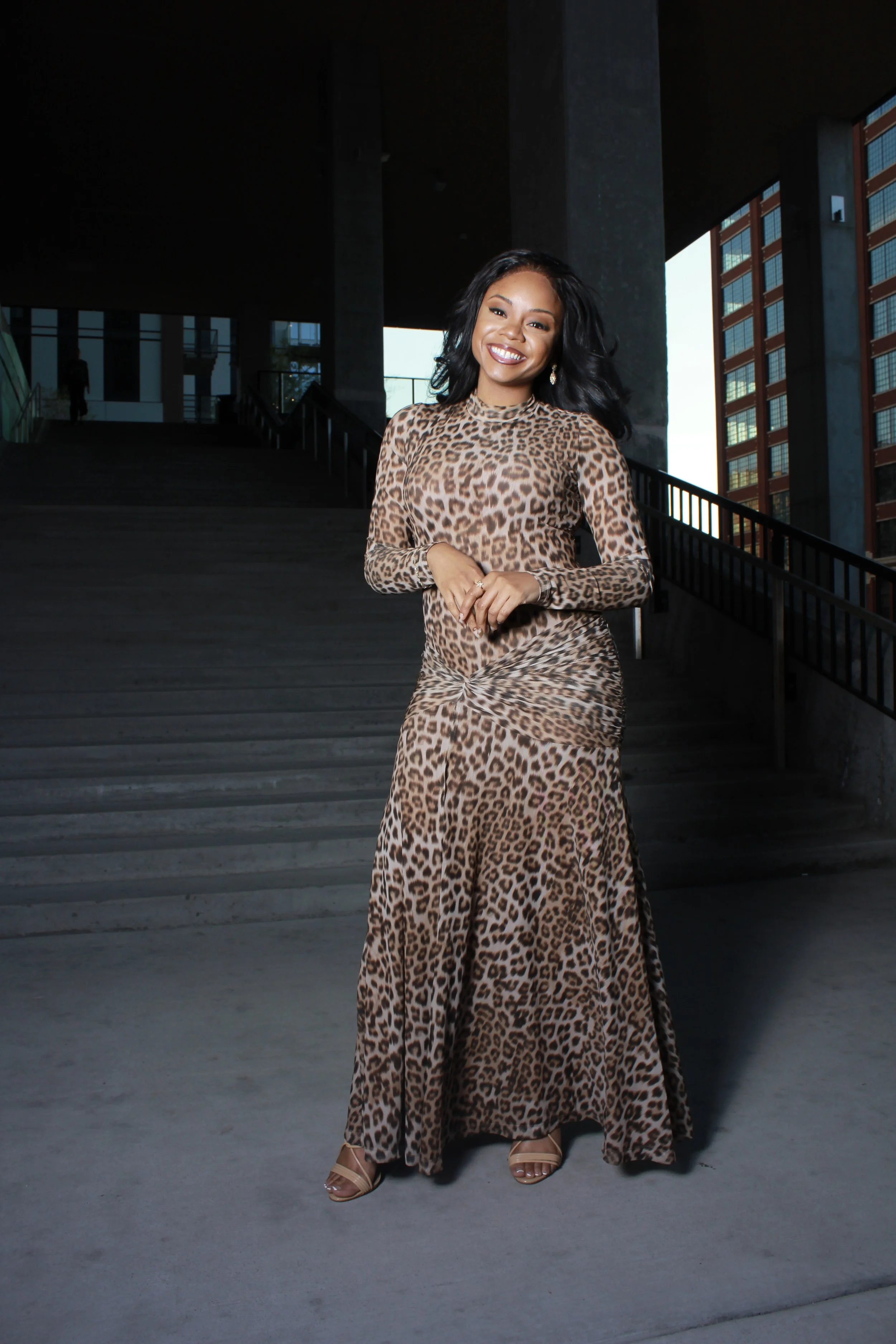 A woman wearing a leopard print long sleeve dress and heels, smiling and standing on steps outside an urban building with large windows and concrete architecture.