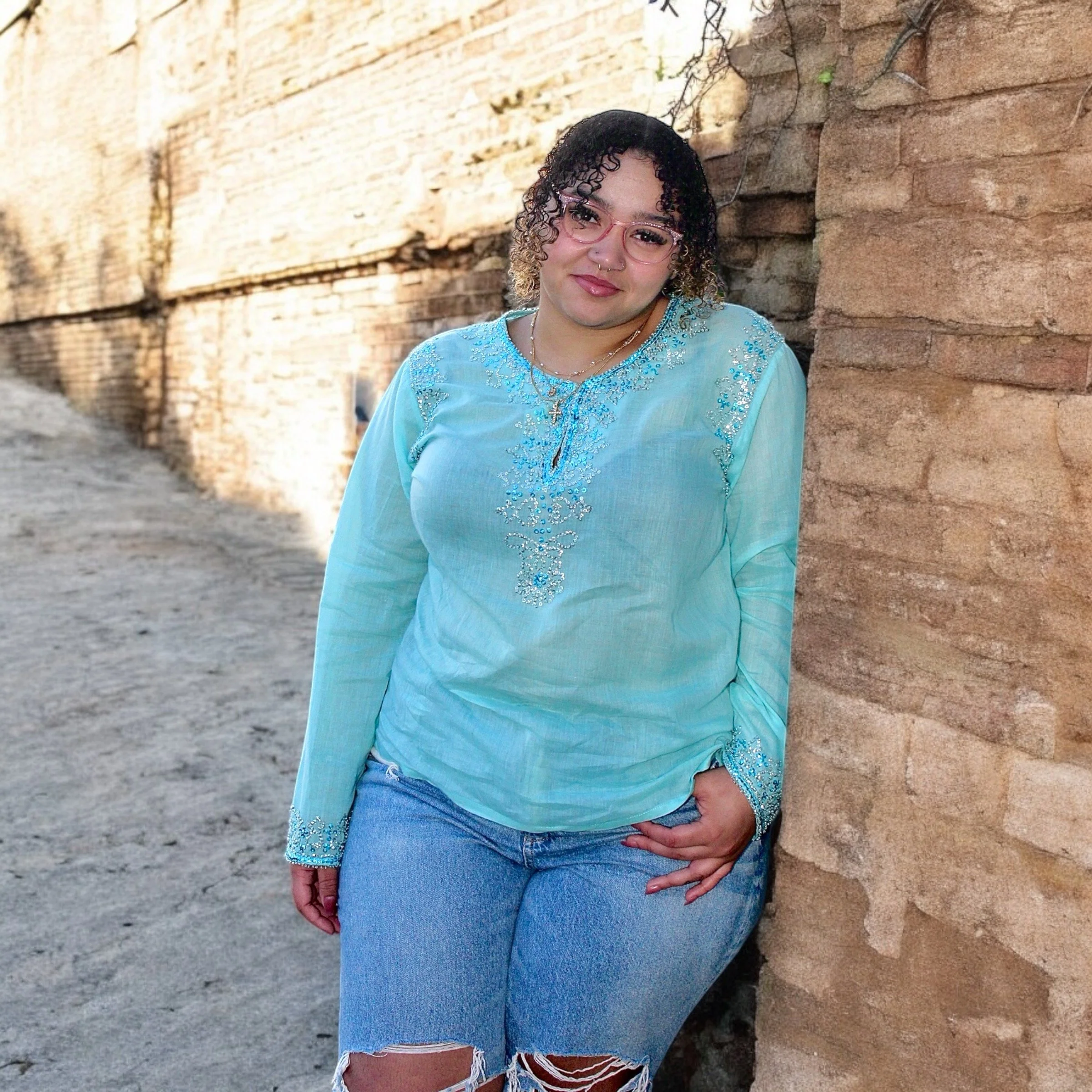 A woman with curly hair, glasses, and jewelry wearing a light blue embroidered top and ripped jeans, standing against a brick wall.