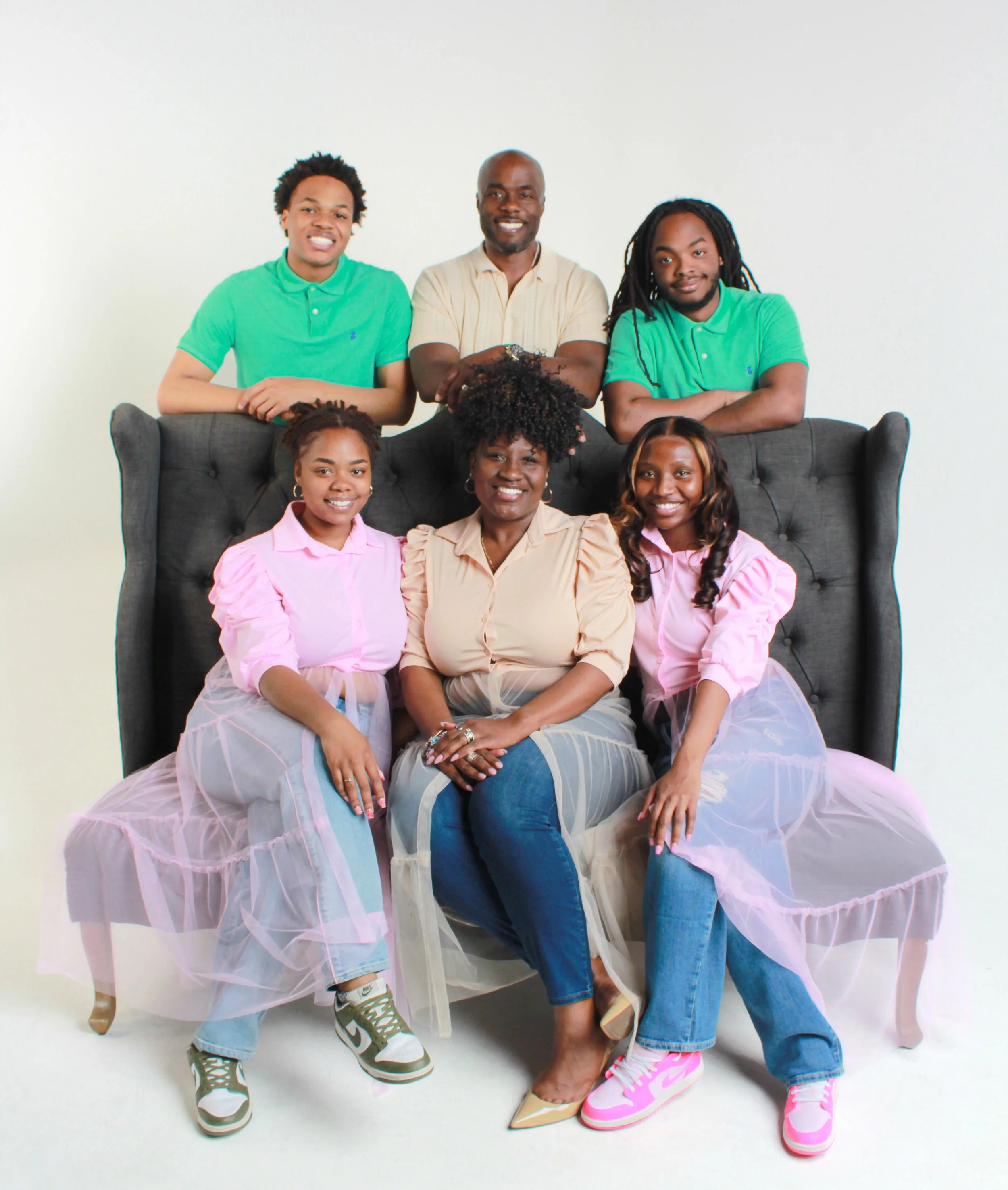 Group of seven people, four women sitting and three men standing behind them, smiling, in front of a plain white background. Women are wearing pastel pink tulle skirts and matching pink sneakers, while men are dressed casually with sneakers.