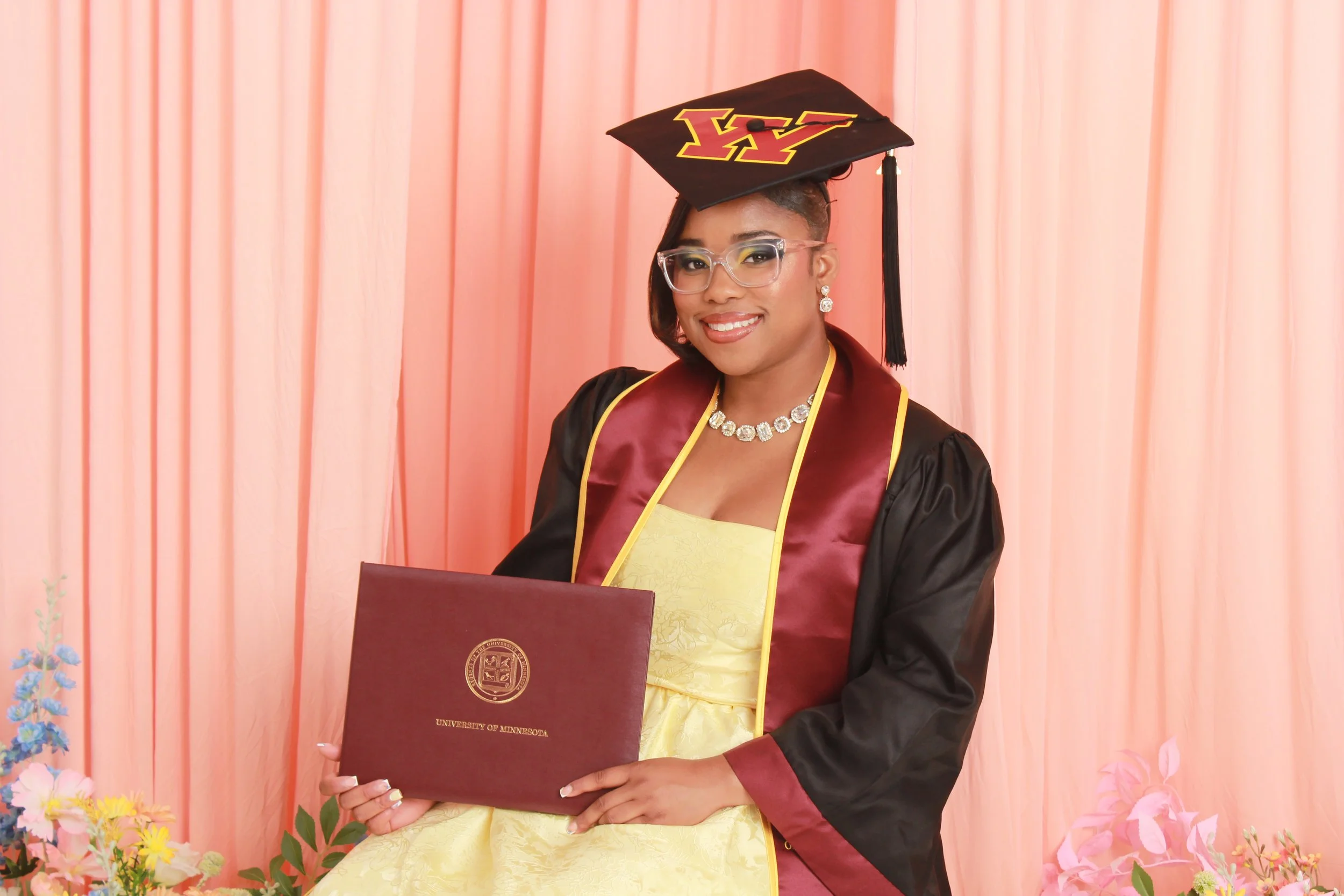 A woman in a graduation cap and gown holding a university diploma in front of a pink curtain backdrop, with flower decorations in the foreground.