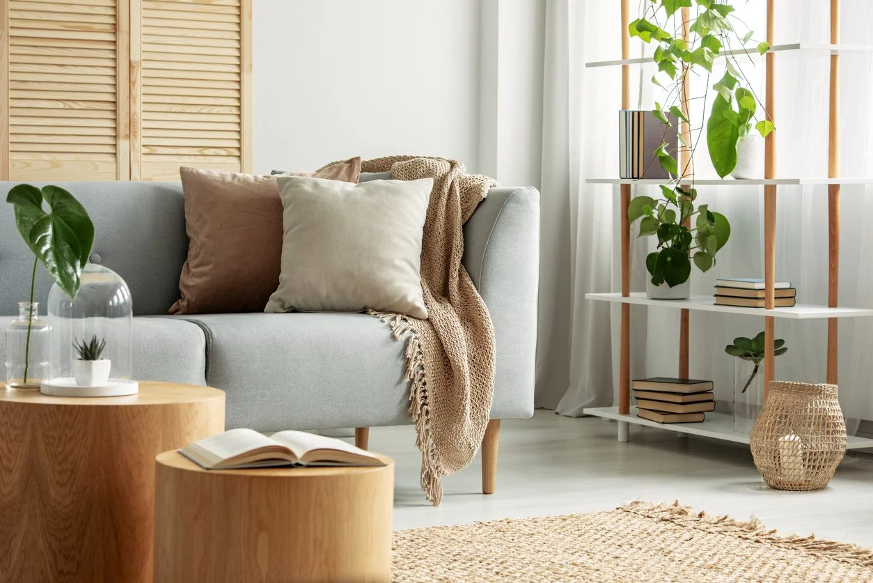 Living room with a light gray sofa decorated with brown and cream pillows and a beige throw blanket. A wooden coffee table with an open book and a smaller matching table are in front. Behind the sofa is a wooden screen, and to the right is a white shelf with potted plants and books. The room has natural light coming through white curtains, creating an airy and cozy atmosphere with neutral and earthy tones.