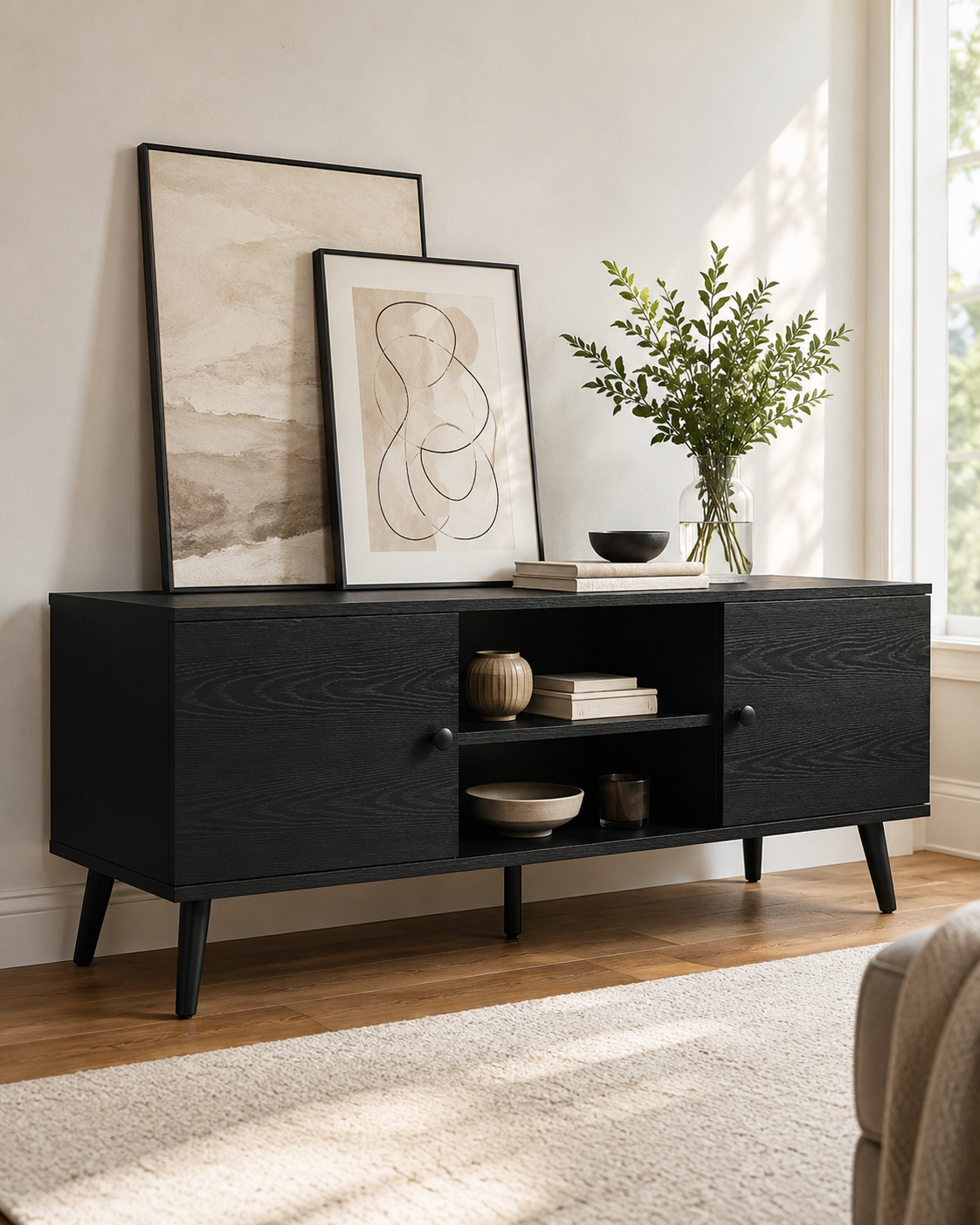 A black wooden sideboard with decorative art frames, a glass vase with greenery, and small decorative items on top, set against a white wall near a window with natural light.