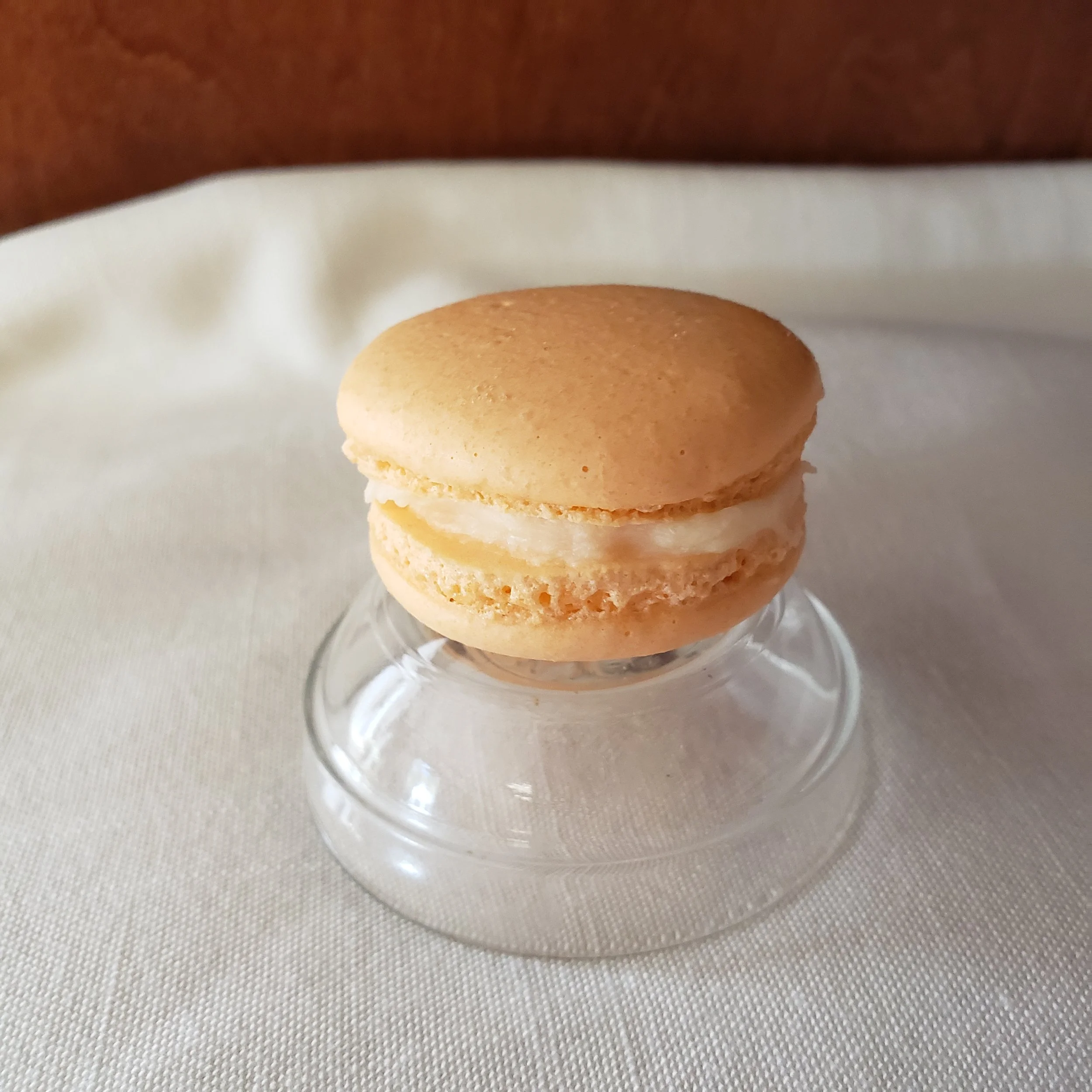 Close-up of a stack of light brown macarons with a white filling, placed on a glass dish with a decorative pattern, on a textured light-colored surface.
