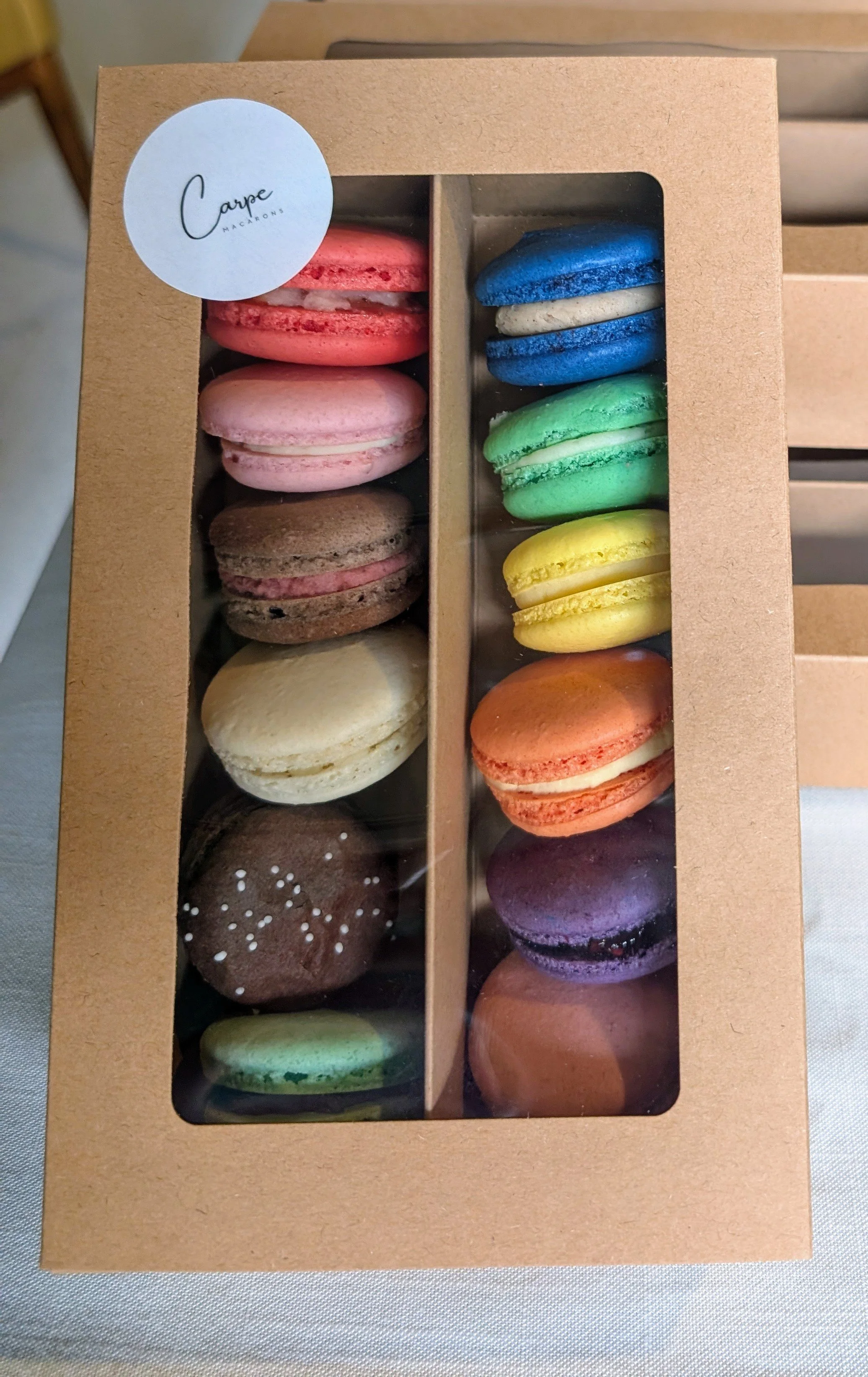 A cardboard box with a clear window containing assorted colorful macarons arranged in two columns.