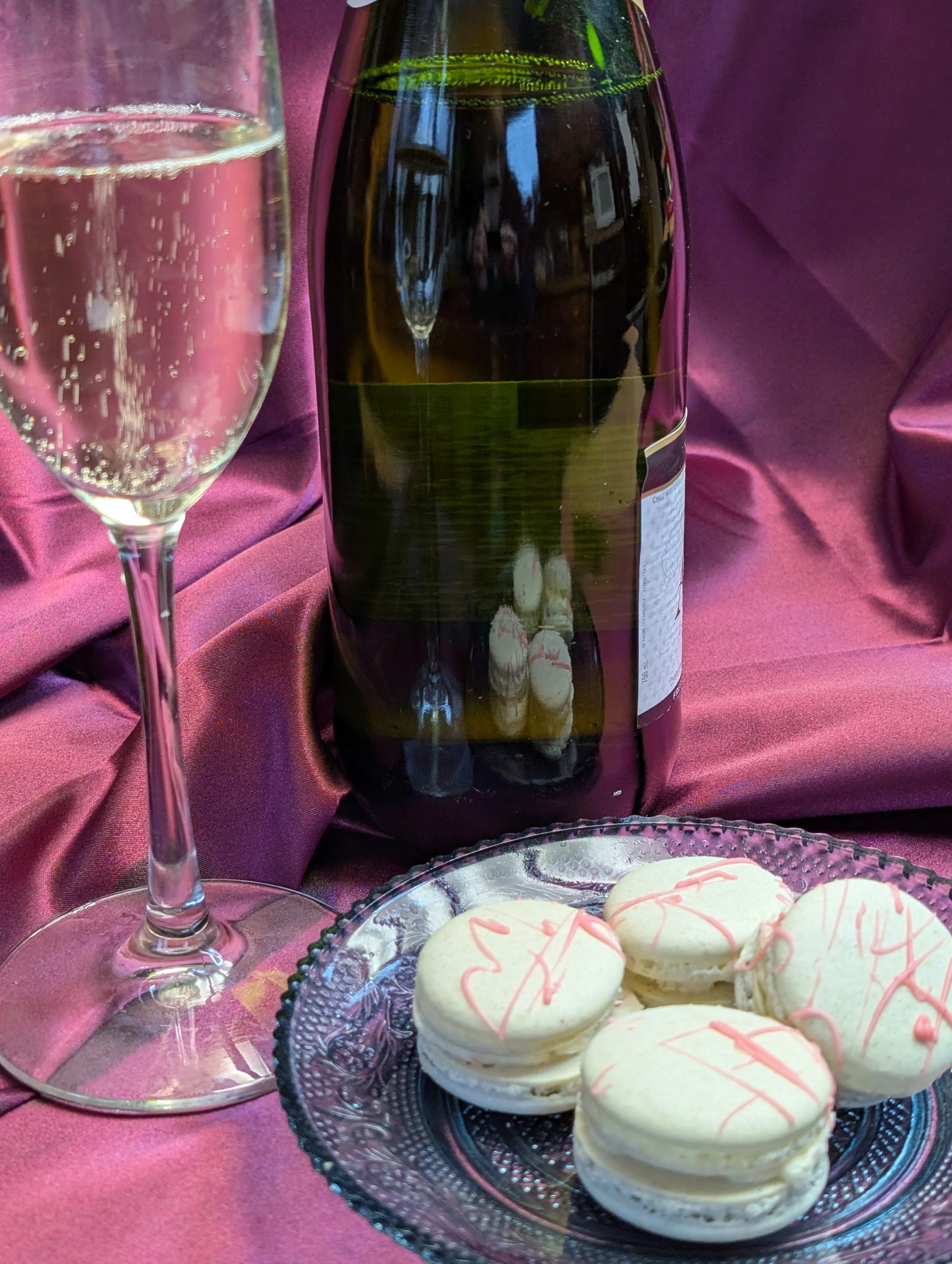 A glass of sparkling wine or champagne, a brown glass bottle, and a plate of white macarons with pink drizzle on a pink satin tablecloth.