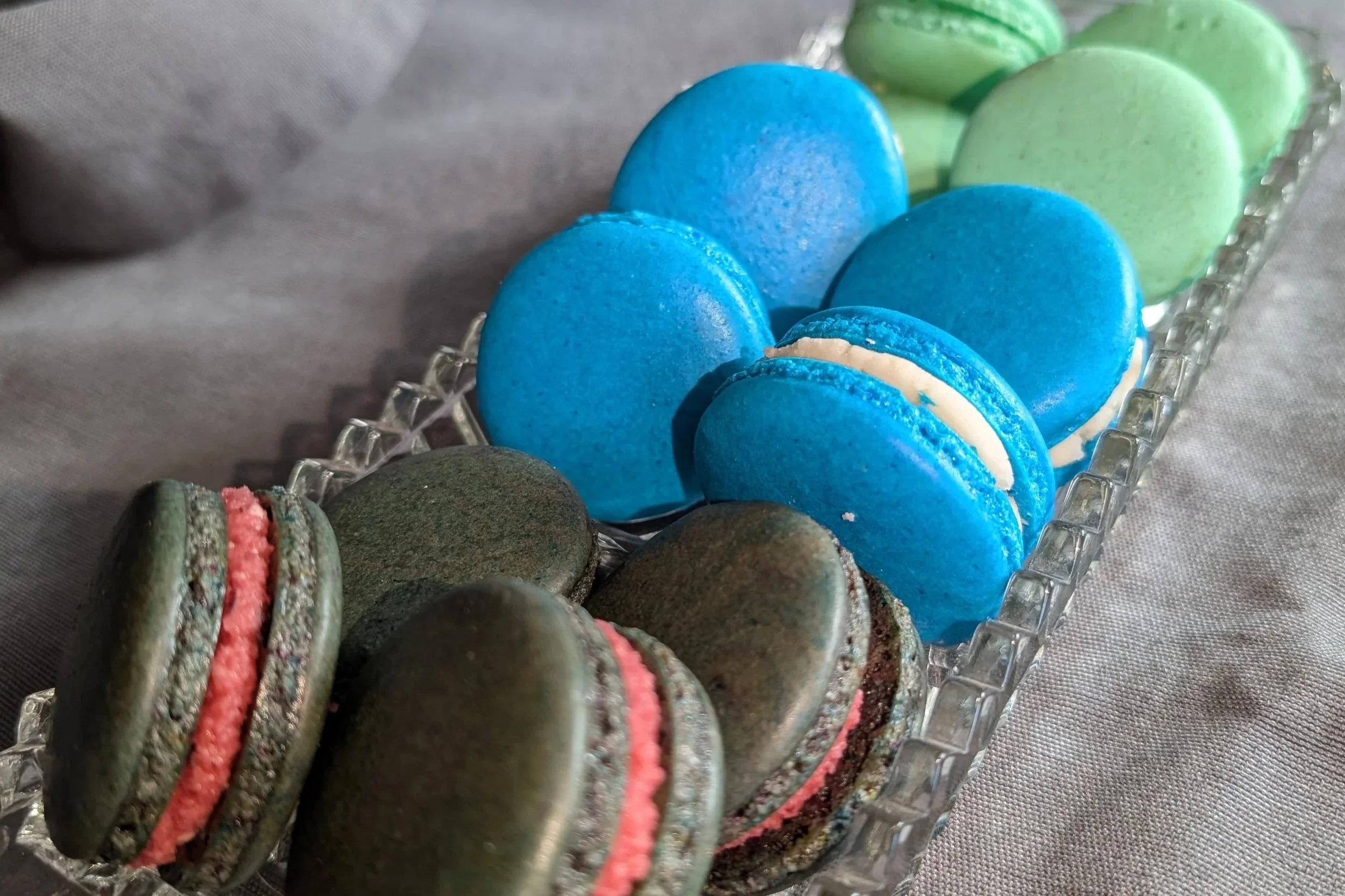 A glass tray with rows of colorful macarons in blue, green, and pink.