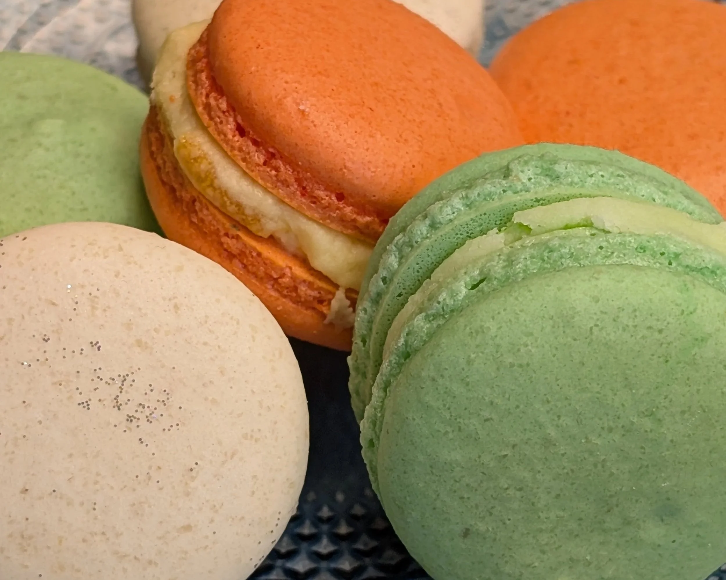 Close-up of various colorful macarons, including green, beige, and orange, on a dark textured surface.