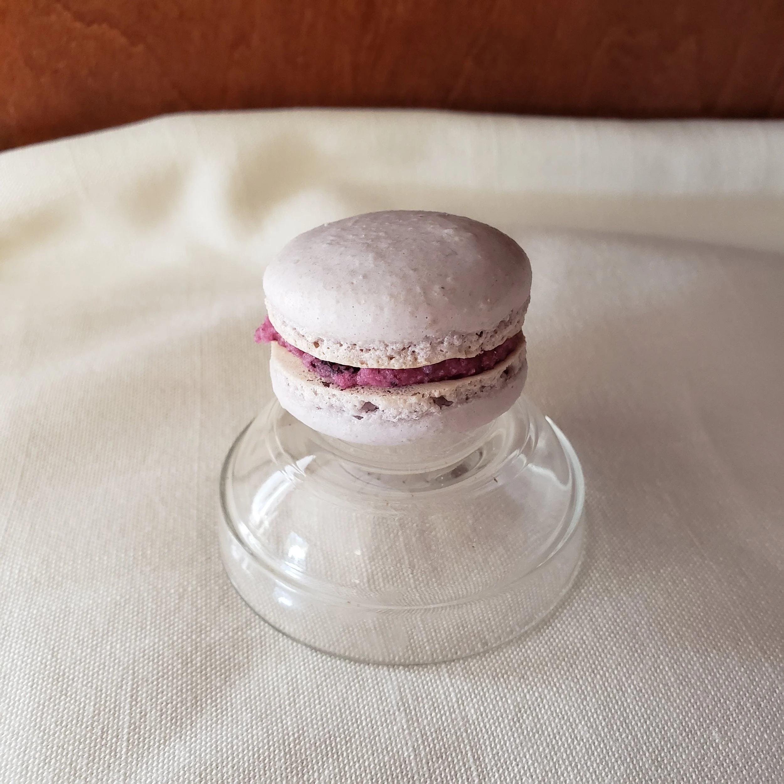 A pink macaron with filling, placed on a glass cake stand on a table.