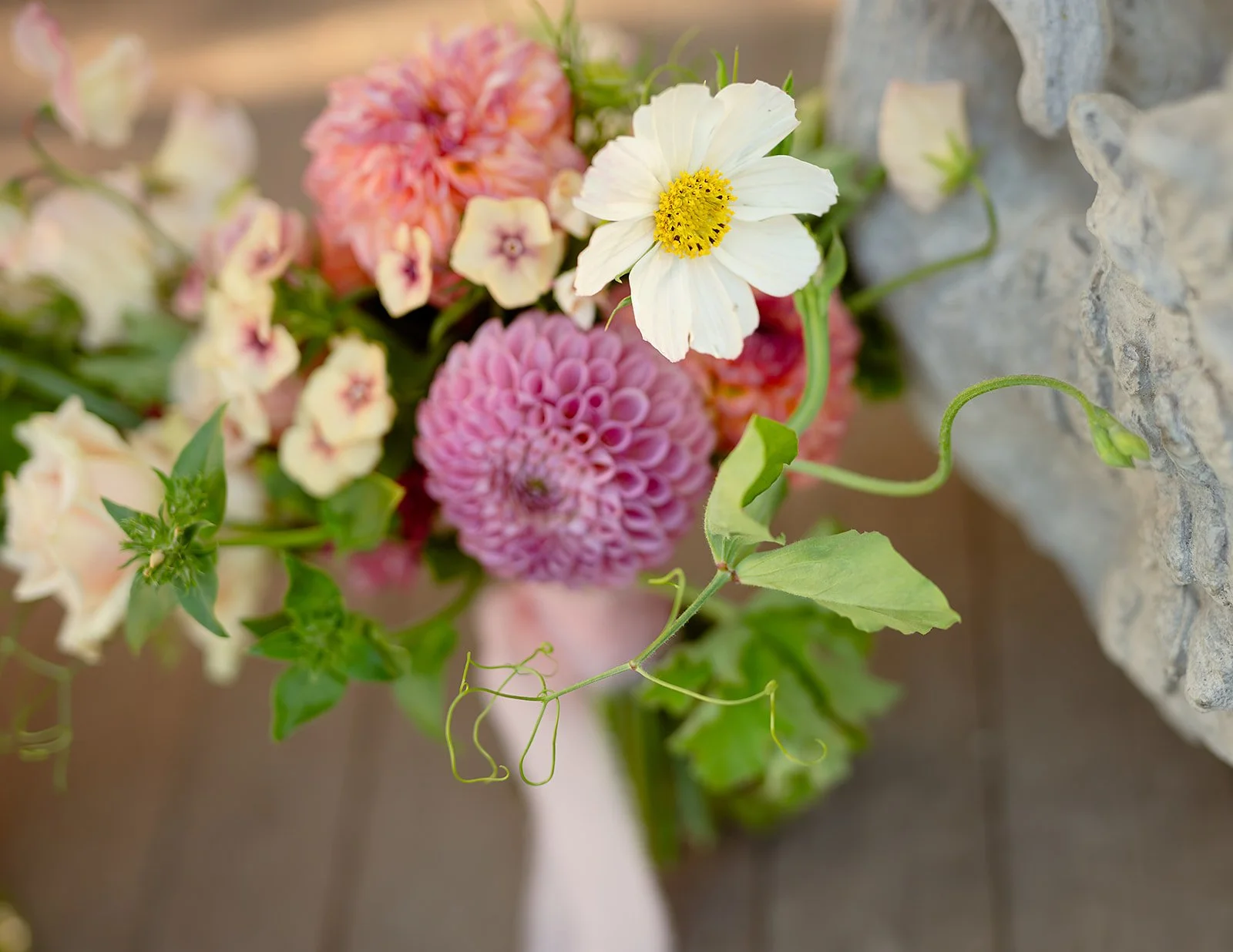 Close-up of a colorful flower arrangement with pink dahlias, white cosmos, small yellow and purple flowers, and green leaves with a stone wall in the background.