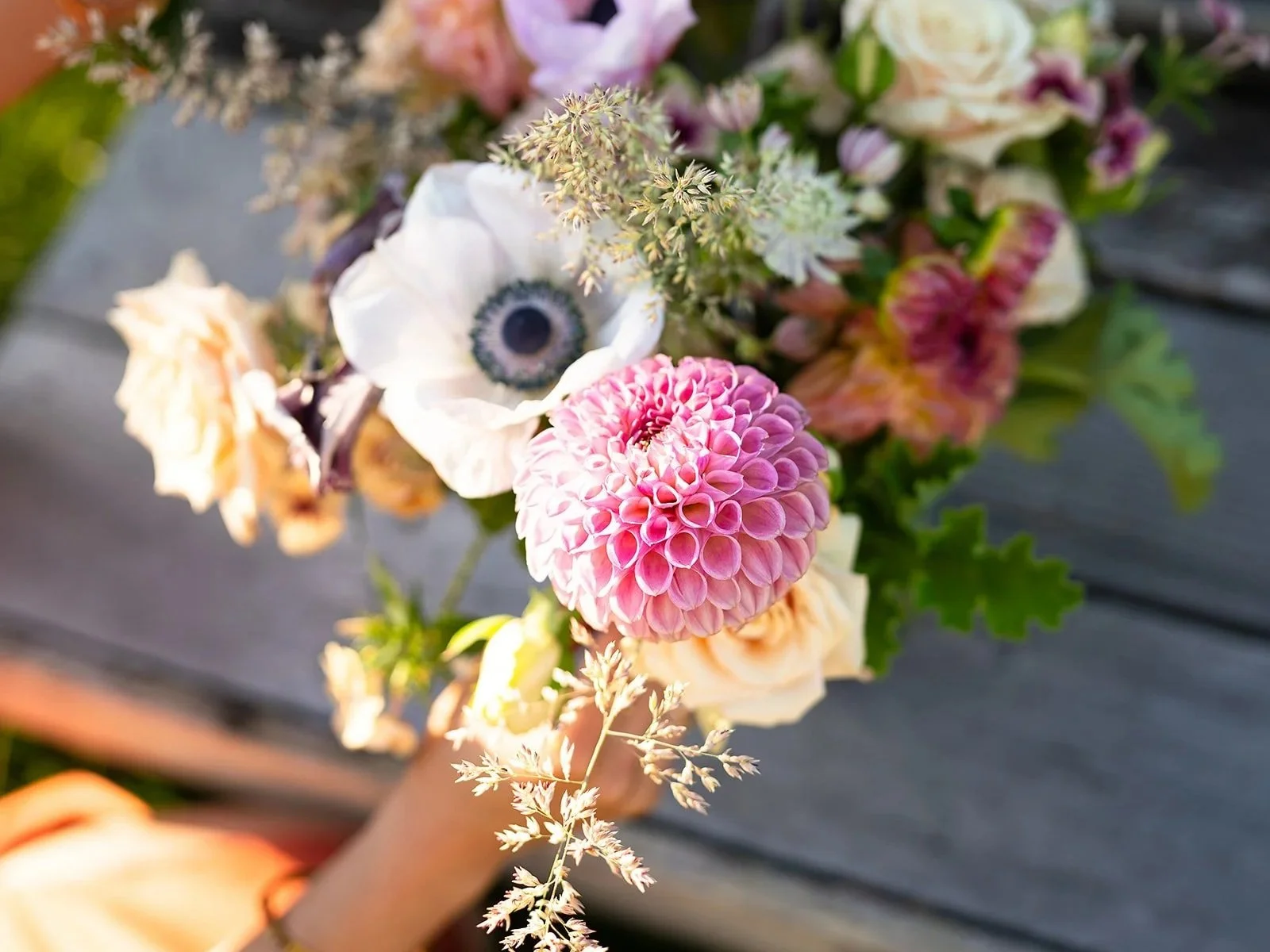 Mixed bouquet of flowers including a tulip, a pink dahlia, a white flower resembling an anemone, and various other blossoms, set against a blurred outdoor background.