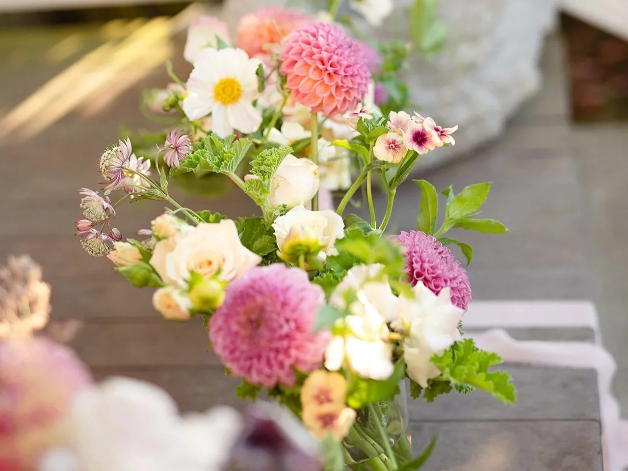 A vibrant bouquet of various flowers including pink dahlias, white and pink primroses, and white roses in a glass vase on a wooden surface.