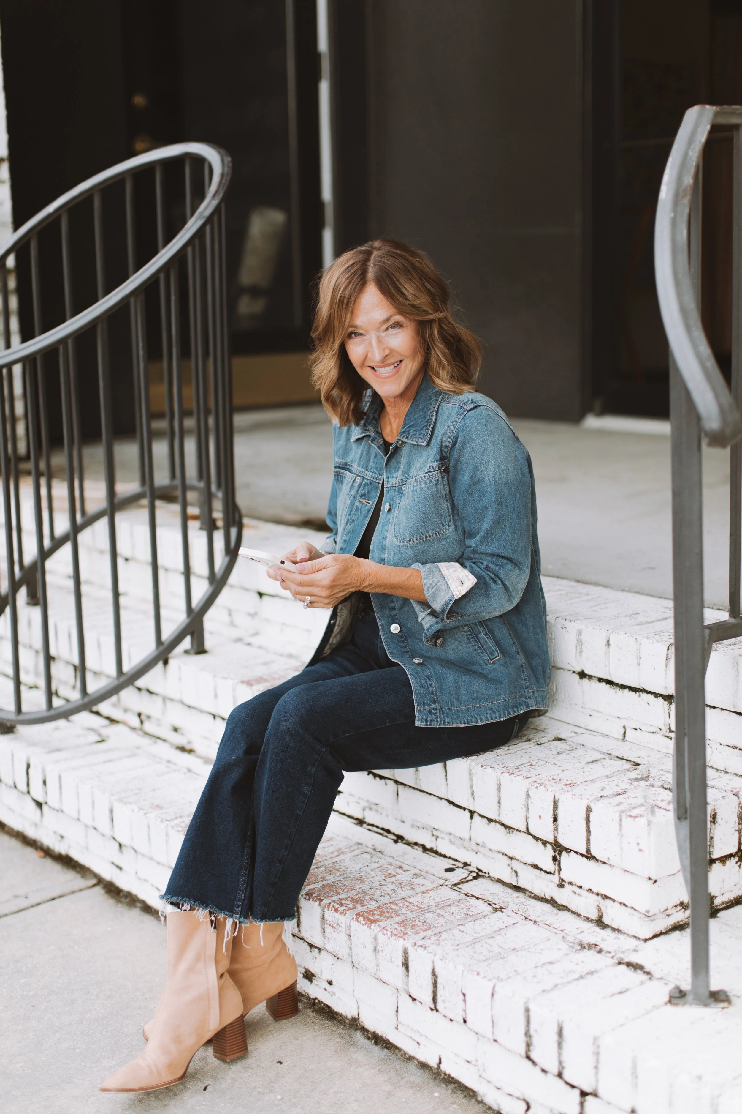 A middle-aged woman with brown hair smiling and looking at the camera, sitting on white brick stairs outside a building, wearing a denim jacket and dark jeans, holding a smartphone, and wearing tan high-heeled boots.