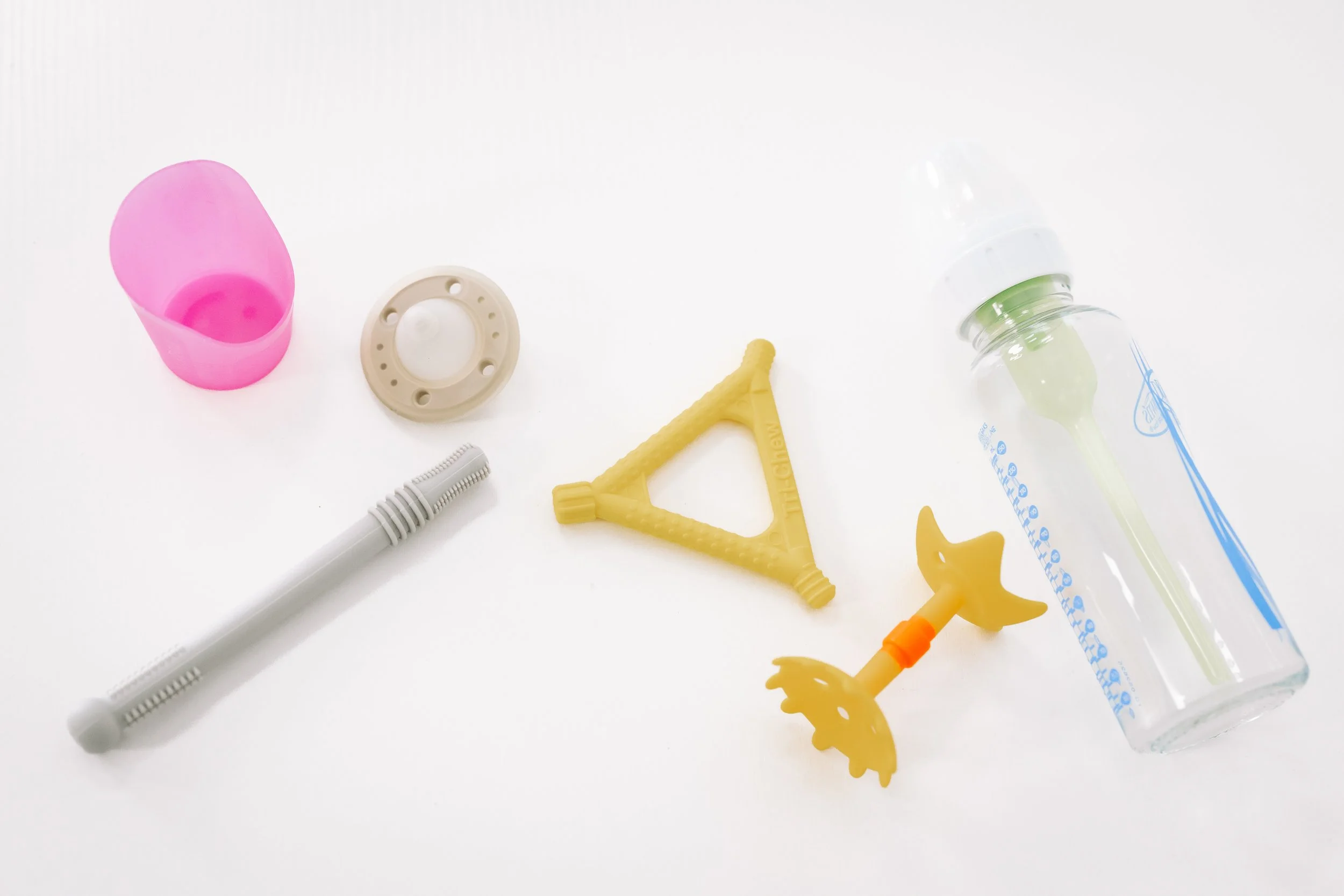 Baby feeding supplies including a pink sippy cup, a beige pacifier, a yellow teething toy, a white and gray straw, and a baby bottle with a green nipple.