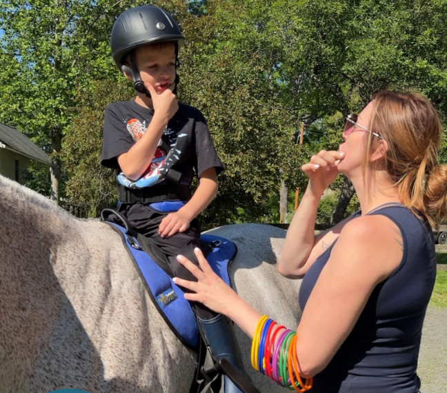 A young boy wearing a helmet sitting on a horse, talking to a woman with sunglasses and colorful bracelets, outdoors with trees in the background.