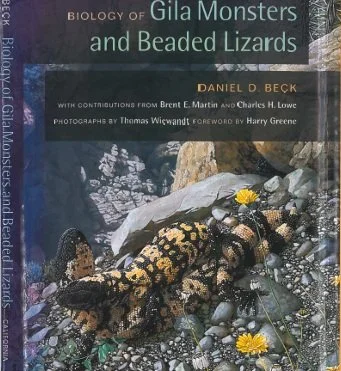 Close-up of a book cover titled "Biology of Gila Monsters and Beaded Lizards" by Daniel D. Beck, featuring a Gila monster with black and yellow scales on a rocky terrain with small yellow flowers.